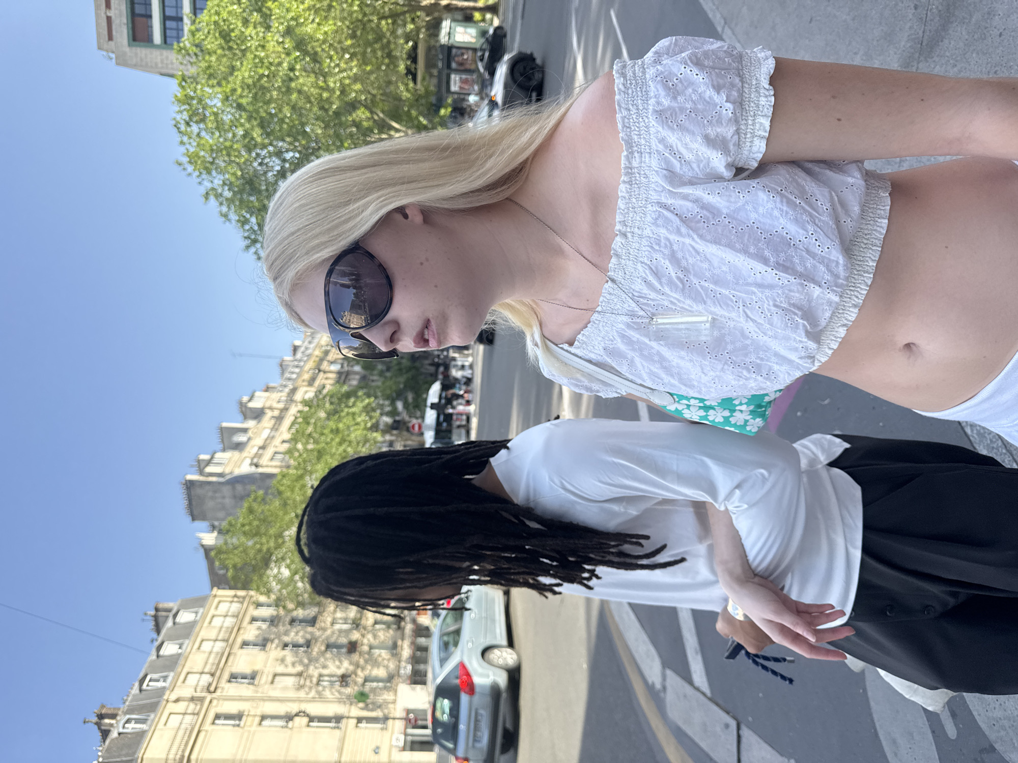 Two individuals are present, likely in Paris, France, based on the architectural style of the surrounding buildings. The person in the foreground is a blonde woman wearing sunglasses and a white eyelet crop top. She has a silver necklace. Behind her is a person with dark dreadlocks, wearing a white long-sleeved shirt and black pants. The street appears to be a crosswalk with cars and scooters visible in the background. The buildings are traditional Parisian structures. The sky is clear and blue.