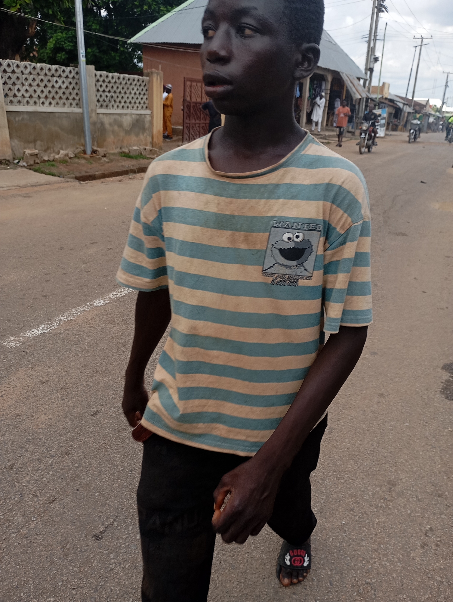 A young Black male walks on a paved road in Shendam, Nigeria. He is wearing a horizontally striped t-shirt with beige and turquoise stripes, featuring a cartoon character graphic on the left chest. He is also wearing black trousers and black sandals with red and white accents. The road is asphalt, with a white line marking the edge of the lane. Buildings with corrugated metal roofs and concrete walls are visible along the roadside, as are utility poles and trees. In the background, several other people and motorcycles are present. The weather appears overcast.