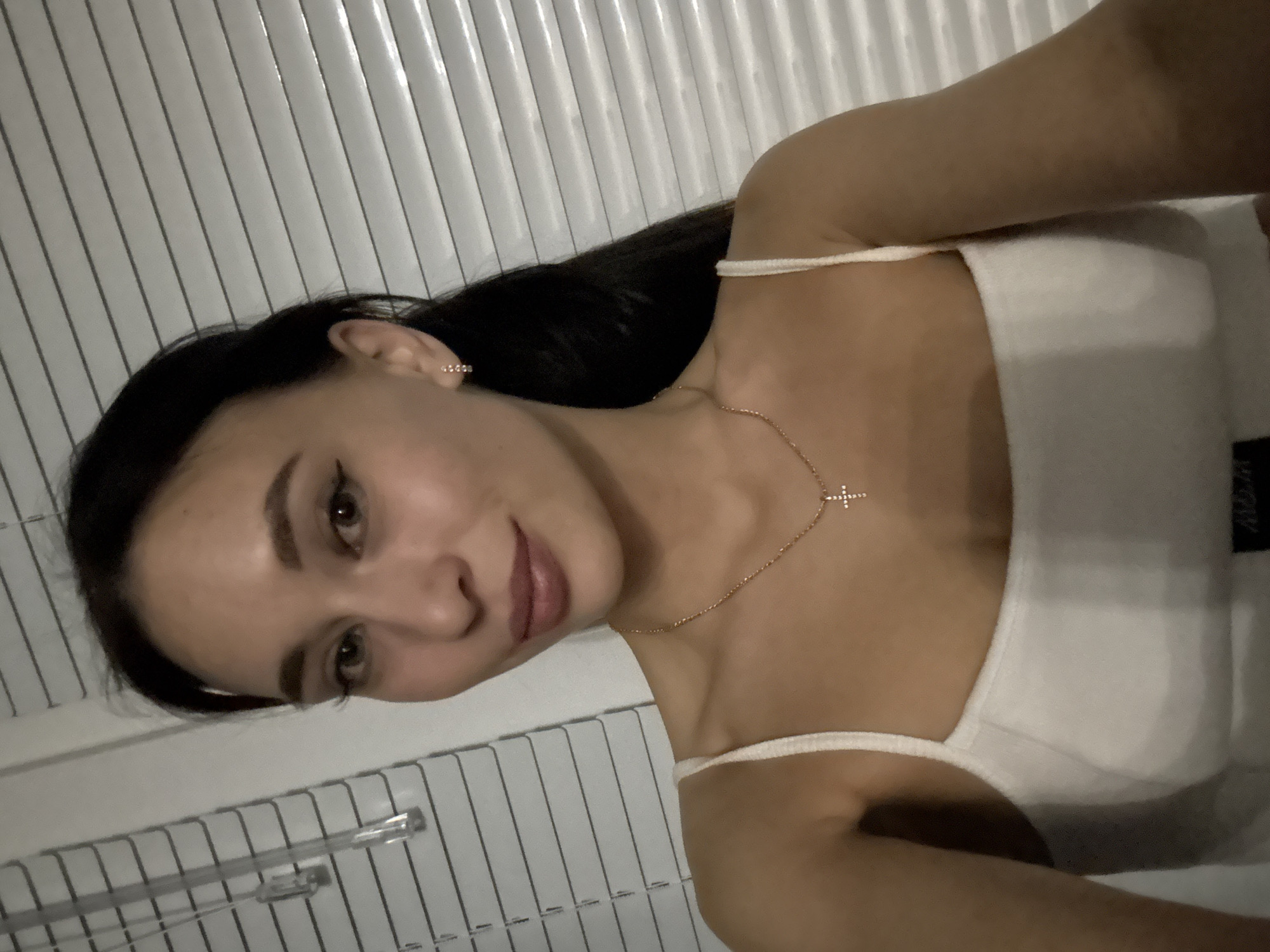 A young woman is photographed from the upper chest up. She is wearing a white tank top and a gold cross necklace. She has dark hair and her expression is neutral. The background is a white Venetian blind. There is no information indicating that the photo was taken in the city of Chaikovsky, Russia.