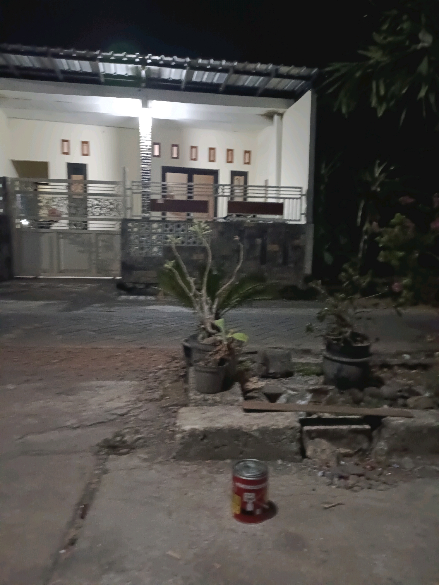 The image depicts a dimly lit outdoor scene at night, likely in Rancang Kencono, Indonesia. A modern house with a prominent roof structure and white walls is visible in the background. The house has several square windows or frames. A decorative metal fence runs across the front of the property, with a gate visible.
In the foreground, on the right side of the image, are two potted plants. The one closer to the viewer is in a dark, cylindrical pot and has sparse, dry branches. The second pot, to its left and slightly further back, also appears to contain a plant with sparse foliage. The ground in front of the house is paved with interlocking bricks and concrete slabs, some of which appear cracked or uneven.
A single red and yellow tin can is placed upright on the concrete ground in the lower-center of the image, casting a slight shadow. Debris, including small stones and what appear to be bits of wood, is scattered around the base of the potted plants and along the edge of the concrete. The overall lighting suggests it is nighttime, with some artificial light illuminating the house. The scene appears quiet and still.