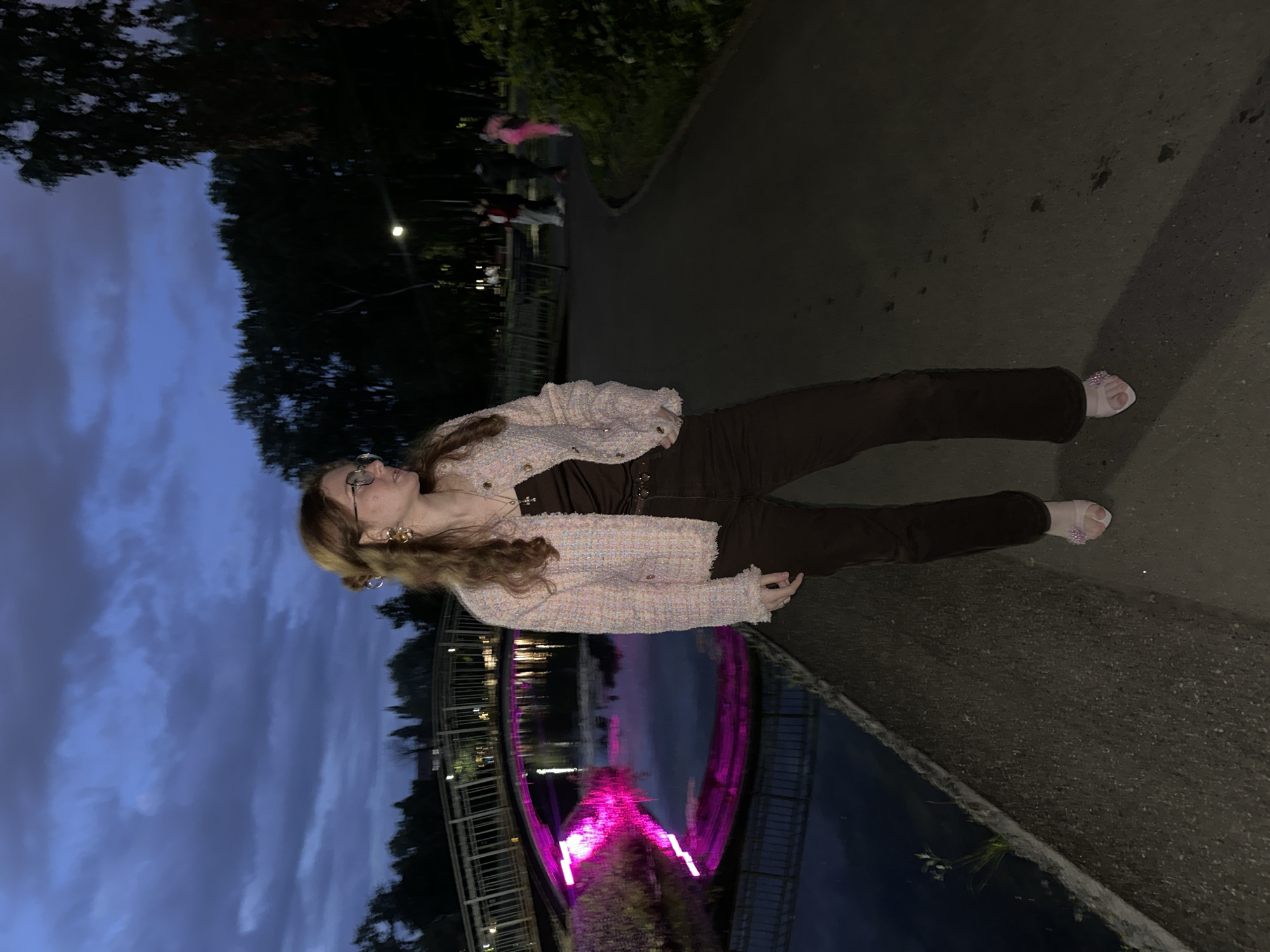 The image depicts a young woman standing on an asphalt path next to a body of water in an urban park in Kazan, Russia, during twilight or night. The specific location is identified as Gorky Park in Kazan, evidenced by the illuminated pedestrian bridge over a waterway.
The primary subject, a woman with light brown, wavy hair tied in a partial bun, wears eyeglasses, large gold-colored earrings, and a necklace with a cross pendant. She is dressed in a light-colored (pink and blue textured fabric) jacket over a dark brown sleeveless top and dark brown trousers. Her footwear consists of light-colored open-toe sandals embellished with a decorative element on the top. She stands facing to the right, with her head turned to profile, looking beyond the frame.
To her left, a calm body of water reflects the dark sky and a prominent bright pink light source. The water is bordered by a concrete retaining wall. Spanning the water is a modern pedestrian bridge, partially illuminated underneath by vibrant pink lighting, which creates a strong reflection. The bridge's structure includes visible railings and a pathway. Buildings and trees are discernible in the distance beyond the bridge.
The background to the right reveals a tree-lined park area. A curved, paved path extends into the distance, where several indistinct human figures are visible. One figure in the background is wearing a bright pink garment, while others are in darker attire. A single streetlamp emits a bright halo among the trees, providing additional illumination to the scene. The sky above is a dark, cloudy blue.