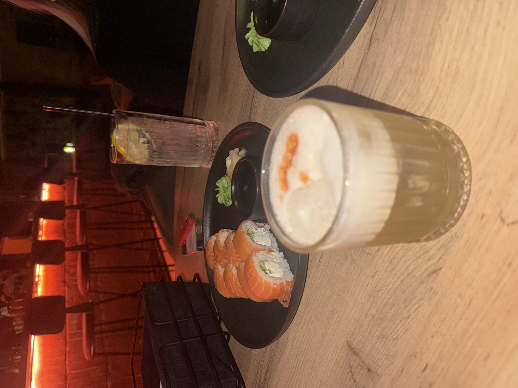 The image shows a tabletop scene with a black plate of sushi rolls, a small container of soy sauce with wasabi, and a cocktail with a foamy top, as well as a drink in a textured glass with a straw. The setting appears to be a restaurant or bar in Batumi, Georgia, as suggested by the chairs and a dimly lit red brick wall in the background. The tabletop is made of wood. There are no people directly visible in the frame. A small red object that appears to be a lighter can be seen near the plate of sushi.