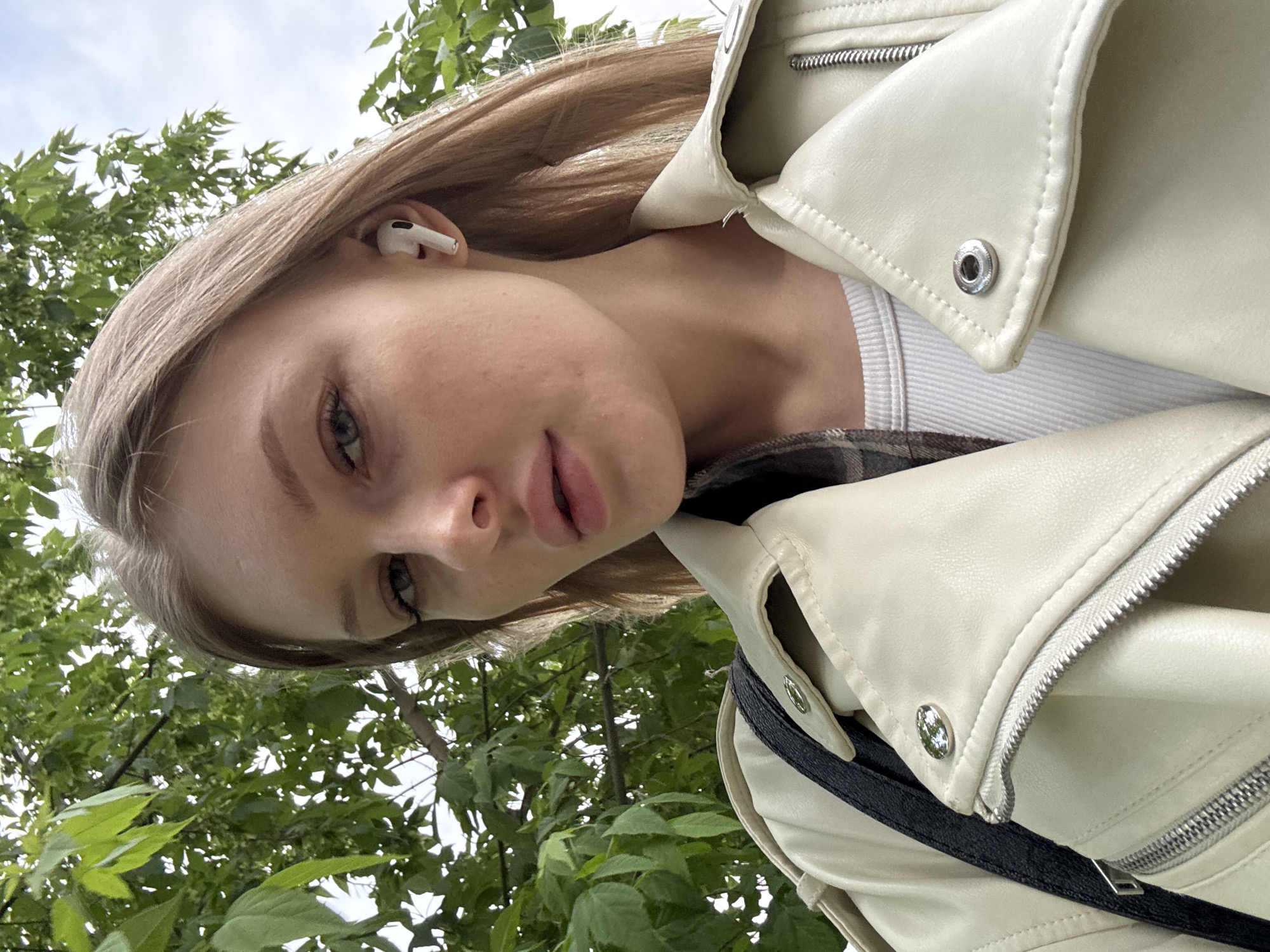 A young woman with light hair and fair skin, wearing a white leather jacket, a white ribbed tank top, and an earbud in her left ear, appears to be taking a selfie in an outdoor setting. The background features green foliage and a glimpse of the sky. Based on analysis, the location is Perm, Russia.