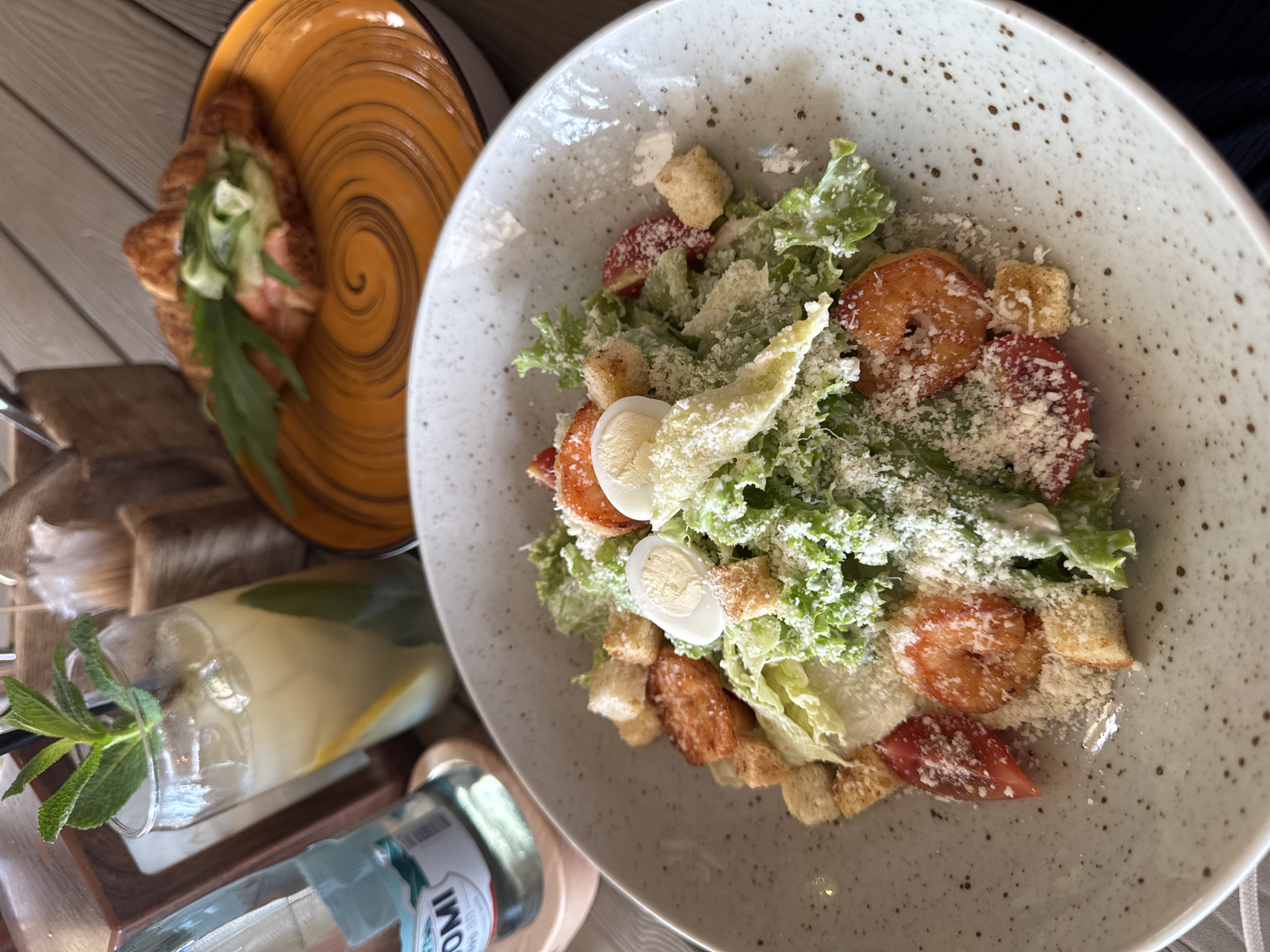 The image shows a table setting with a Caesar salad with shrimp and quail eggs in a speckled grey bowl. Next to it is a plate with what appears to be a croissant sandwich filled with salmon and greens. A glass bottle of lemonade with mint is visible near the salad. The scene is likely a restaurant or cafe setting, but the specific location in Sirius, Russia, cannot be determined from the image alone. No people are visible.