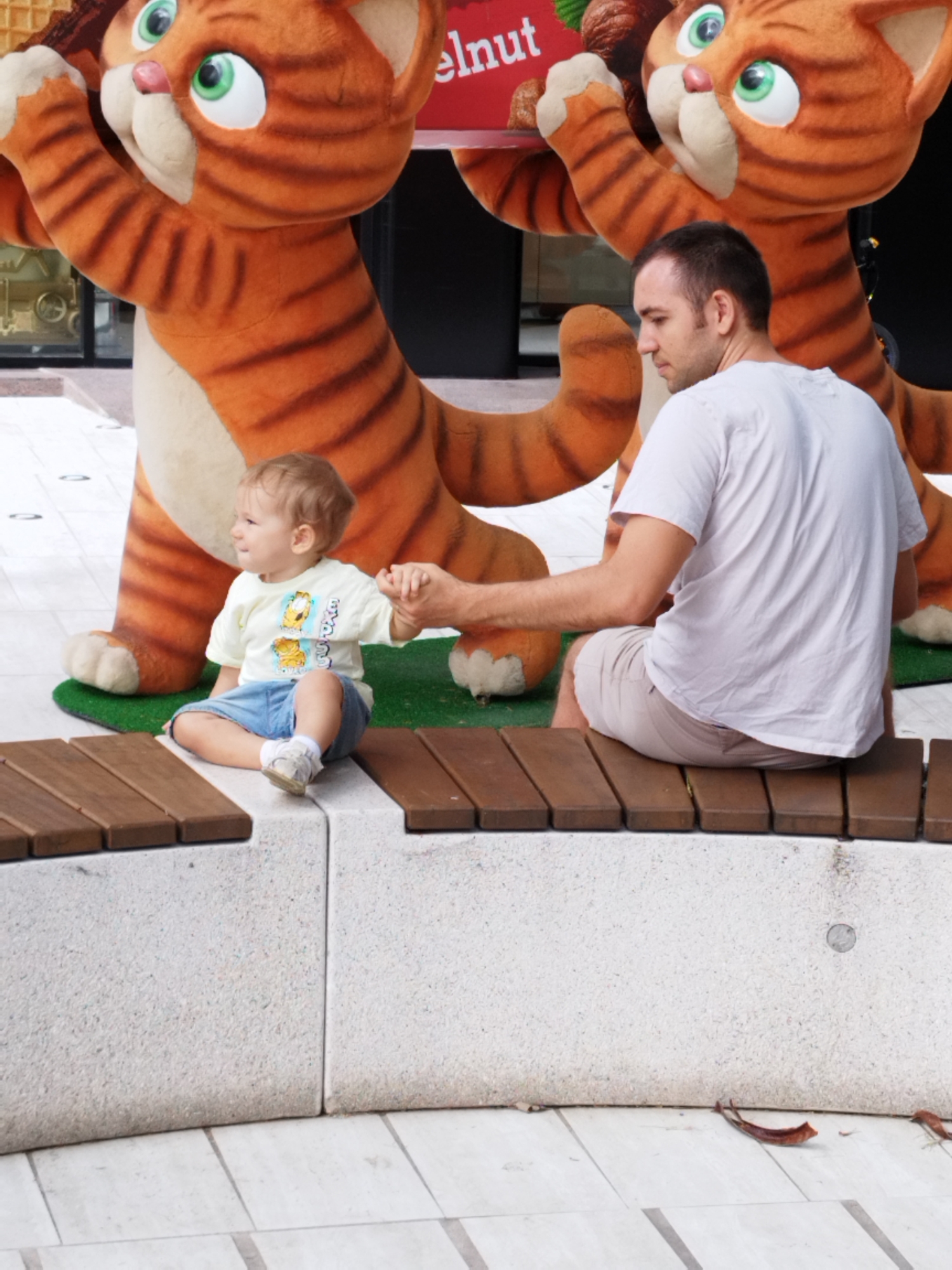 A man and a child are sitting on a stone bench. The child, a toddler, is sitting on the bench, wearing a light yellow t-shirt with cartoon characters and blue shorts. The man is sitting beside the child, wearing a white t-shirt and light-colored shorts. The man is holding the child's hand. In the background, there are large, plush cat figures with orange and white stripes and green eyes. The cat figures appear to be part of a promotional display or decoration. The environment appears to be an outdoor public space with a paved floor and a stone bench with wooden slats on top. Based on the context provided by the original prompt, this scene is located in Bucharest, Romania.