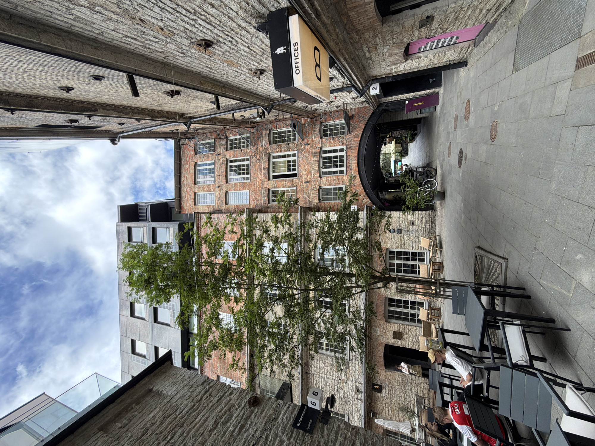 The image depicts a narrow cobblestone alleyway in Tallinn, Estonia, framed by old brick and stone buildings. A modern building with dark cladding and glass windows stands tall on the left, contrasting with the historic architecture. A mature tree with green foliage is centrally located, reaching towards the sky. Outdoor seating with dark metal tables and chairs is arranged on the left side of the alley, with several people seated. A sign with the word "OFFICES" is mounted on the right wall. The alley continues into the distance, leading to an arched passageway with more shops and seating visible beyond. The sky is partly cloudy.