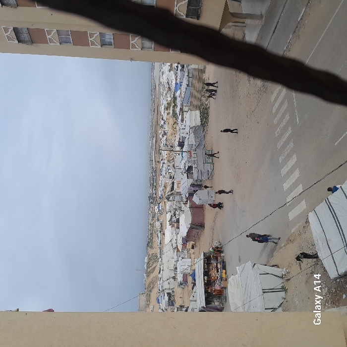 A high-angle, slightly tilted view reveals a sprawling settlement of tents and makeshift structures, possibly a refugee camp or informal urban area. A road with zebra crossings cuts through the settlement, with several individuals walking along it. Above, a building with distinctive window designs frames the top of the shot. A grey sky indicates overcast conditions. The "Galaxy A14" watermark is visible at the bottom.