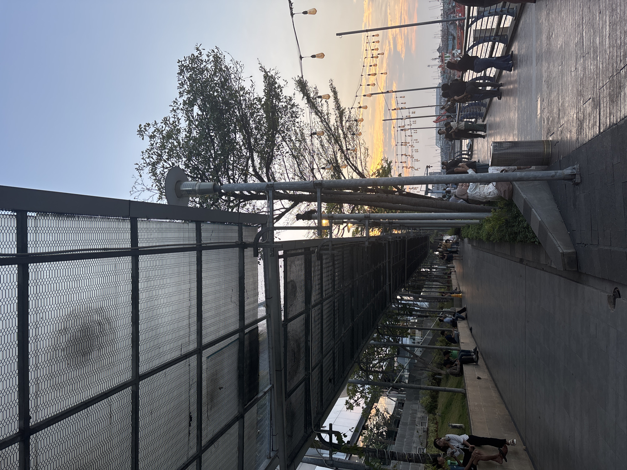 The image depicts a waterfront promenade in North Jakarta, Indonesia, during a sunset. A long, covered walkway with a metal mesh roof stretches into the distance, providing shade over a paved path. Several people are visible, some sitting on a low wall bordering the walkway, while others stroll along the promenade or stand near the water. The setting sun casts a warm glow, illuminating the sky with shades of orange and yellow. String lights are hung along poles on the waterfront, adding to the ambiance. Beyond the promenade, boats are moored in the harbor. The overall scene suggests a leisurely evening at a popular public space.