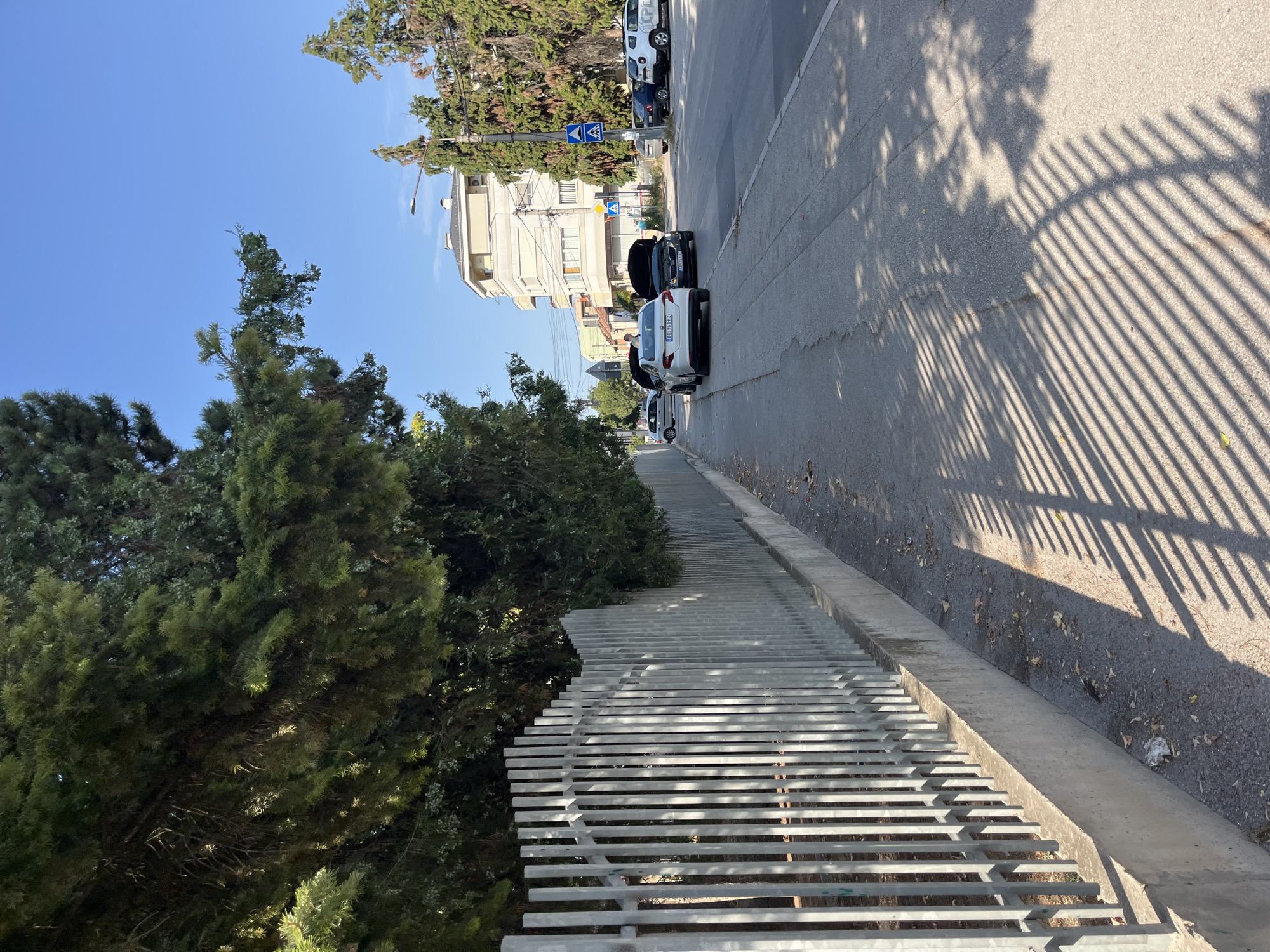 The image depicts a street scene in Čačak, Serbia, on a sunny day. A metal fence lines the left side of the sidewalk, casting a striped shadow across the asphalt. Lush green trees overhang the fence and sidewalk. Several cars are parked along the right side of the street, with two cars in the foreground having their hoods open. One of these cars, a white sedan, is a Fiat. The other, a darker sedan, appears to be a Skoda. In the background, there are multi-story buildings and more trees. Pedestrian crossing signs are visible on the right side of the road.