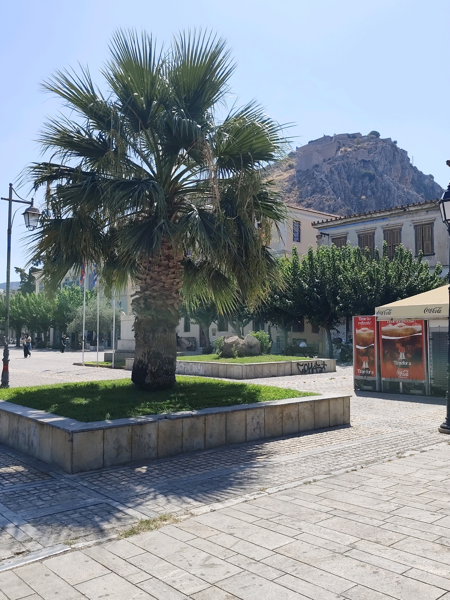 The image depicts a public square in the Municipal Unit of Nafplio, Greece, characterized by a prominent palm tree in the foreground. The square is paved with interlocking stones, and a raised planter box with grass surrounds the base of the palm tree. In the background, a fortified hill, likely a castle or citadel, is visible against a clear blue sky. Several buildings with traditional architecture line the square, some featuring wooden shutters. To the right, there are Coca-Cola advertising displays under a branded awning, suggesting a commercial area. A lamppost is visible to the left. The scene appears to be set on a sunny day, with shadows cast across the paving stones. There are a few people walking in the square, but they are not the primary focus of the image. The overall impression is that of a well-maintained urban space with historical elements.