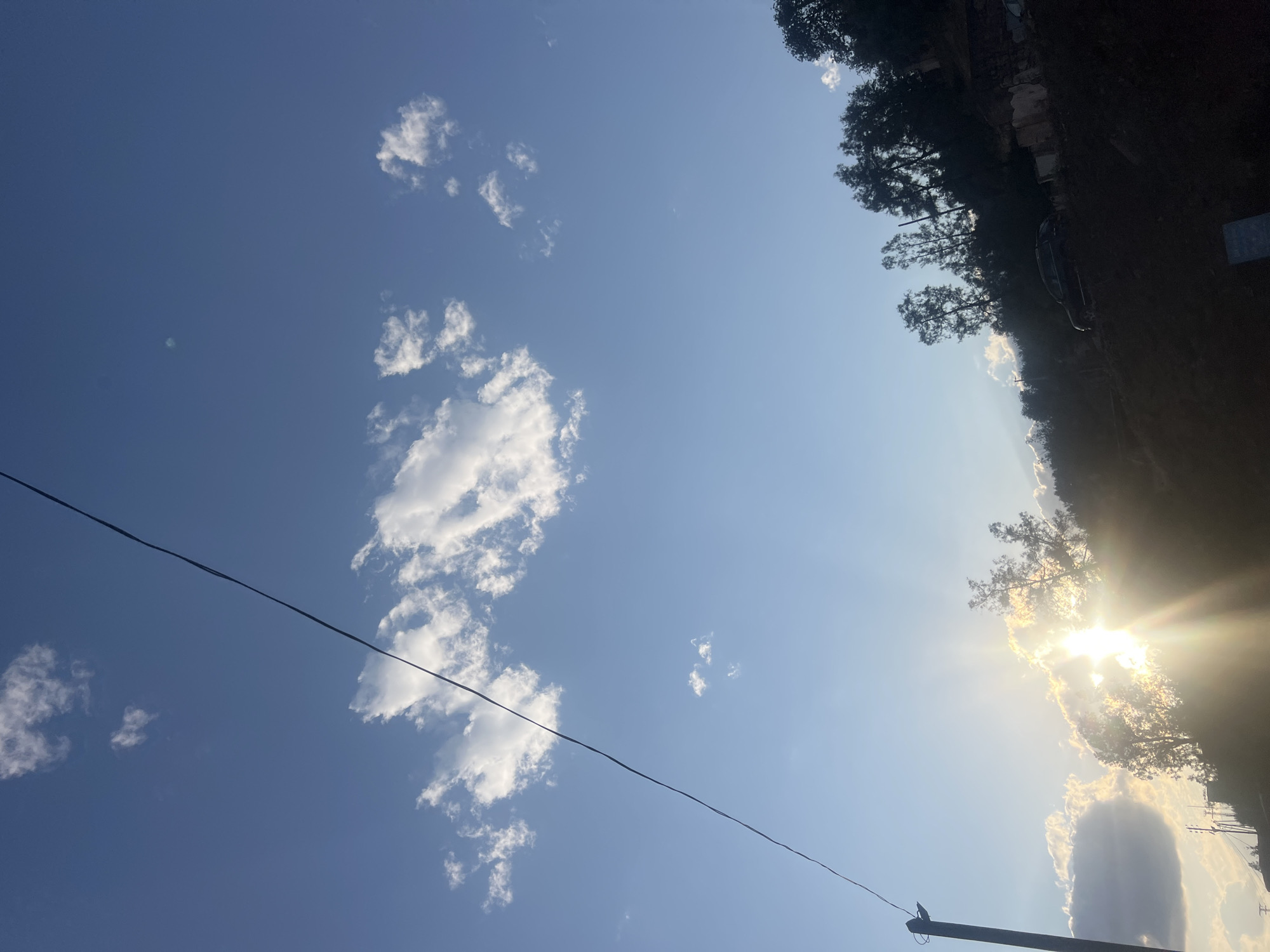The image shows a bright, sunny sky with scattered cumulus clouds. An electrical wire stretches diagonally across the lower-left portion of the frame. On the right, a tree line obscures the horizon where the sun is shining through. A utility pole stands at the bottom of the frame, with a possible bird perched on top. Due to the available information and details in the image, no specific location can be determined.