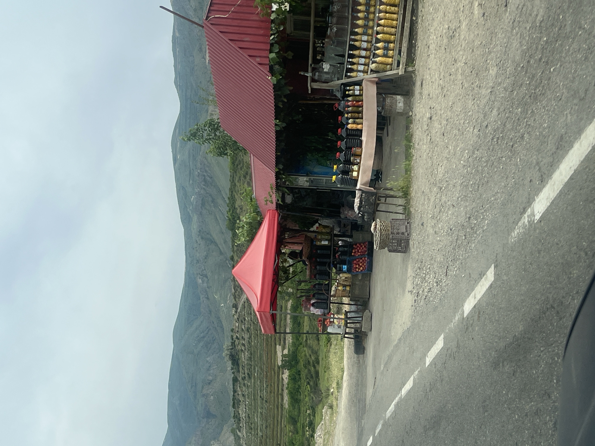 This image depicts a vibrant roadside stall or small market in Chiva, Armenia, set against a majestic backdrop of rugged, terraced mountains under an overcast sky. The photograph appears to be taken from a vehicle, with a paved road and a white dashed line visible in the foreground on the right.
The central subject is a multi-part market structure. On the left, a building with a red corrugated metal roof houses part of the display. Adjacent to this, a bright red triangular canopy extends over an array of goods. The stalls are laden with various products: numerous bottles, mostly amber or yellow in color, are neatly arranged on wooden shelves, suggesting preserved foods, fruit preserves, or possibly local beverages. Below these, larger dark plastic containers are also displayed, likely holding liquids such as oil or honey. Under the red canopy, crates are filled with fresh produce, notably red apples, alongside other fruits or vegetables. Wicker baskets are also visible near the display.
The surrounding landscape features lush green vegetation, with trees near the stall and extensive terraced agriculture climbing the mountain slopes in the middle ground. The time of day is daytime, with an overcast or hazy sky. No people are visible, but the well-stocked display indicates an active trading post. There is no discernible text on any signs or products.