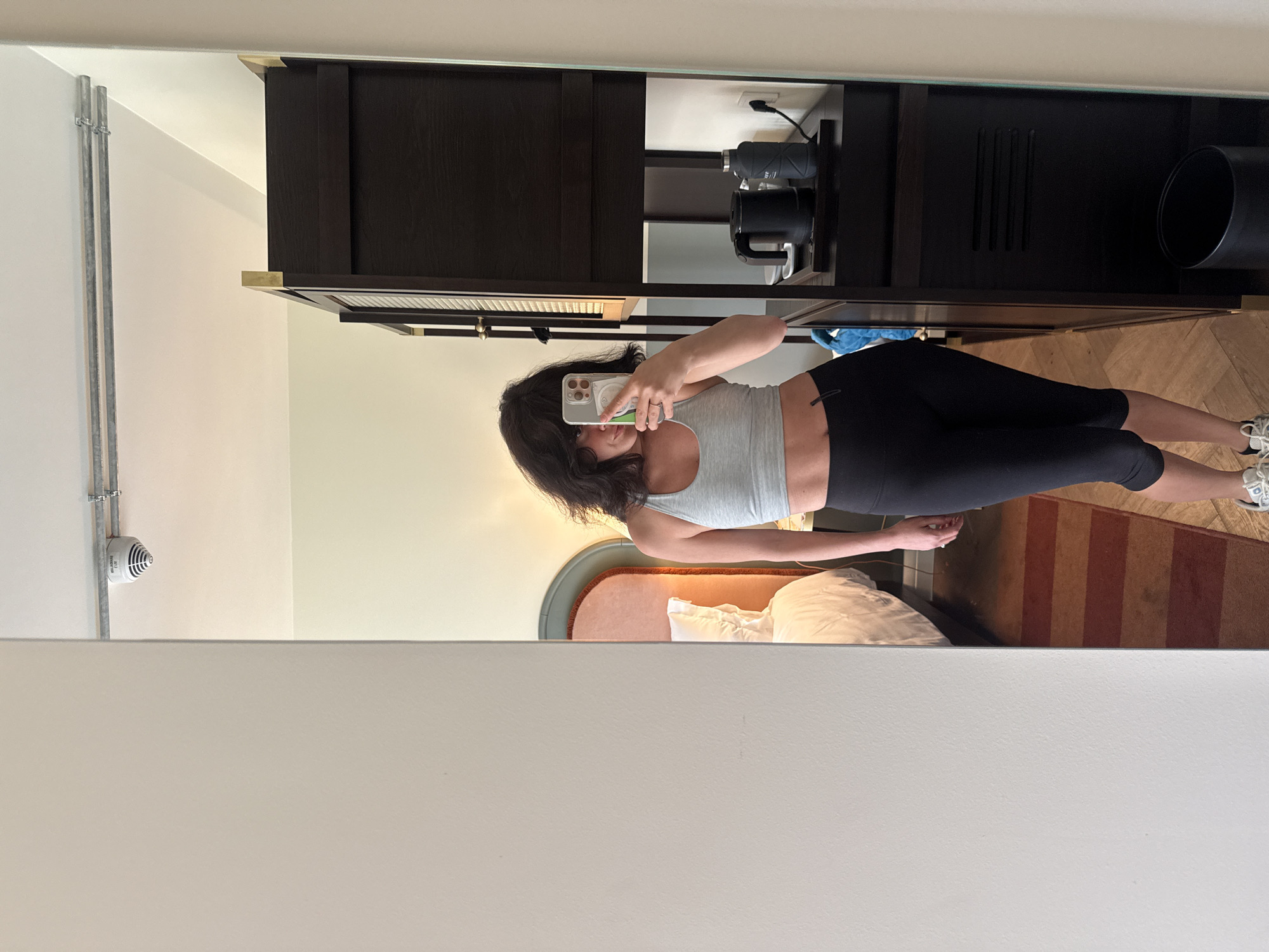 A mirror selfie taken inside a hotel room. The room contains a bed with a salmon-colored headboard, white linens, a dark brown wooden cabinet unit, a black trash can, a coffee maker and water bottle on a shelf. The floor is covered with a striped rug. The person taking the selfie is wearing a light grey tank top and black leggings. A fire safety device is visible on the wall. The location cannot be determined as Berlin, Germany.