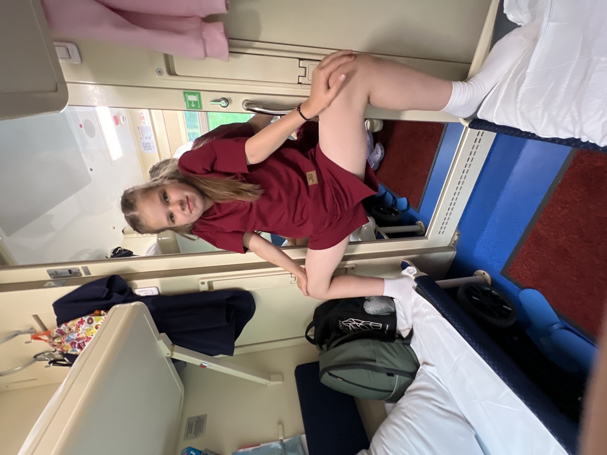 A young girl with blonde hair braided into two pigtails is positioned in a narrow train compartment, leaning forward and resting her elbow on her knee. She is wearing a short-sleeved, dark red t-shirt and white socks. Her expression is a neutral gaze towards the camera.
The compartment appears to be a sleeping berth on a train, with bunks visible above and to the side. The walls are a neutral beige color, with metal trim and door handles. A pink garment is draped over the upper bunk. On the lower bunk, a blue bag and a green backpack are placed. White linens cover the sleeping areas. There is a blue plastic seat in the corner. The floor is carpeted with a dark red material and has a blue accent strip.
The overall scene suggests a travel or a stopover in a train compartment. The lighting appears to be artificial, typical of an indoor train environment, making it difficult to determine the time of day or weather outside. There is no visible text in the image.