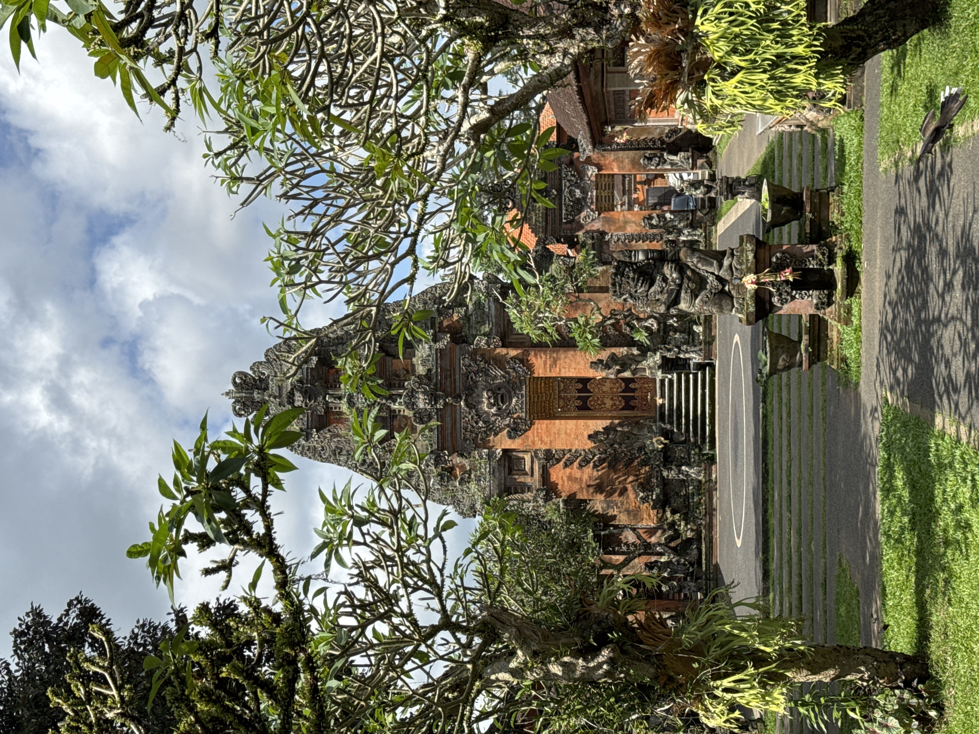 The image depicts a Balinese temple entrance in Ubud District, Indonesia. The temple is constructed of red brick and features intricate stone carvings. A prominent stone guardian statue stands before the entrance, flanked by potted plants. The pathway leading to the temple is lined with grass and stone steps. Lush green trees with bare branches frame the scene, with their foliage partially obscuring the temple's upper structure and the cloudy sky above. The overall setting is serene and traditional, characteristic of Balinese religious architecture.