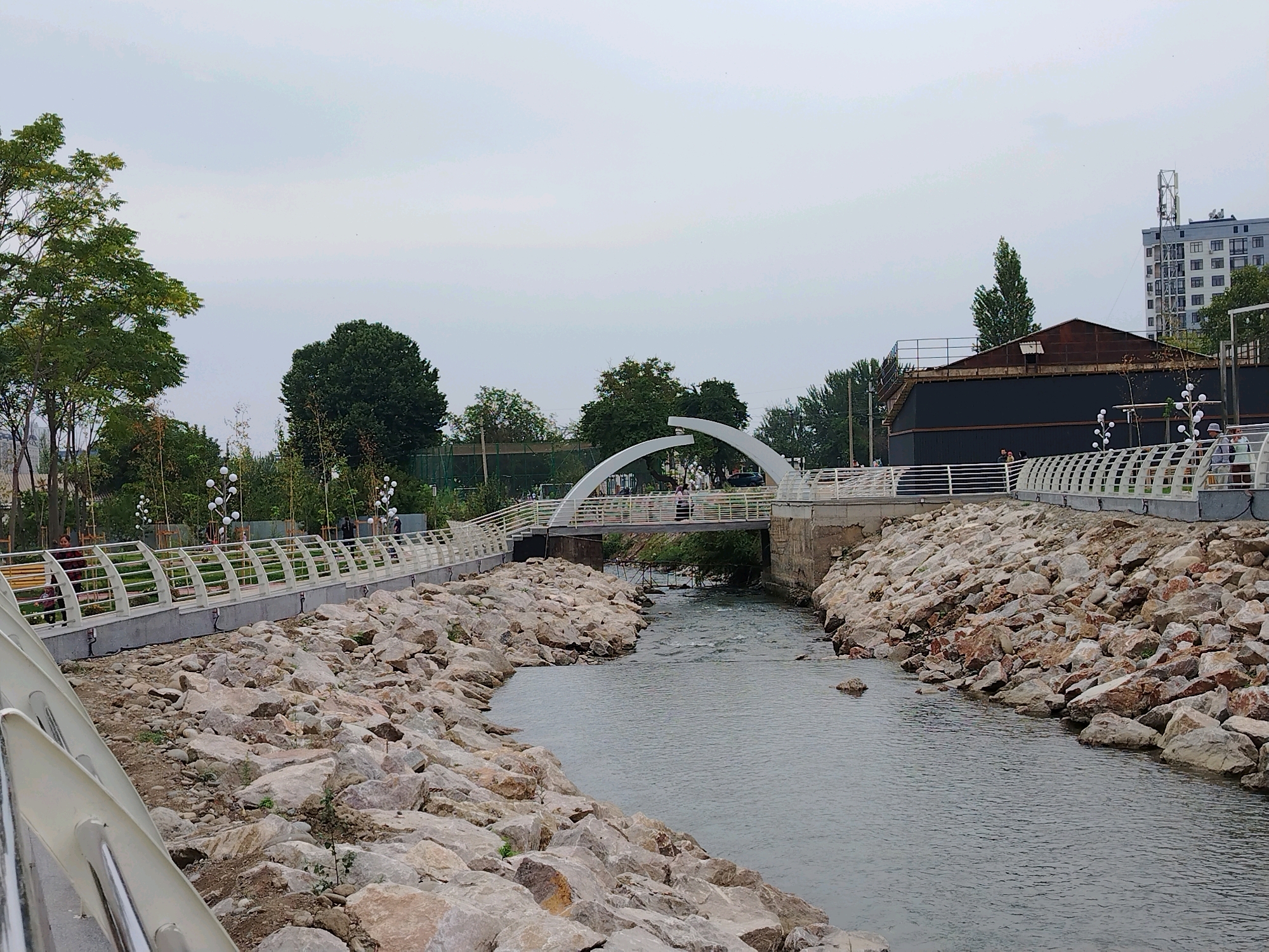 The image shows a waterfront promenade in Osh City, Kyrgyzstan. A river flows through a channel lined with large rocks on both sides. A white arched pedestrian bridge spans the river in the middle distance. Along the riverbanks, there are paved walkways with white railings, some decorative lighting fixtures, and trees. In the background, there is a building with a cell tower and an industrial-looking structure. Several people are visible on the bridge and along the walkways, seemingly enjoying the outdoor space. The sky is overcast.