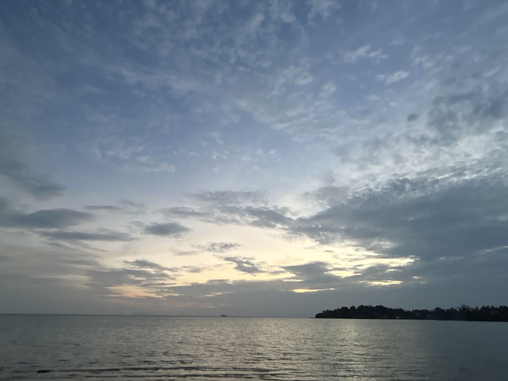 The image depicts a tranquil seascape with a partially cloudy sky at dusk. The water is calm, with subtle ripples reflecting the muted tones of the sky. A faint glow of sunset is visible on the horizon, casting a soft light through gaps in the clouds. To the right, a silhouetted shoreline of trees and land is visible. In the distance, a small, dark shape, possibly a boat or buoy, is barely discernible on the water. The scene is devoid of any people or distinct man-made structures besides the distant silhouette. The overall atmosphere is serene and peaceful, characteristic of a quiet evening by the coast in the Ko Pha-ngan District, Thailand.
