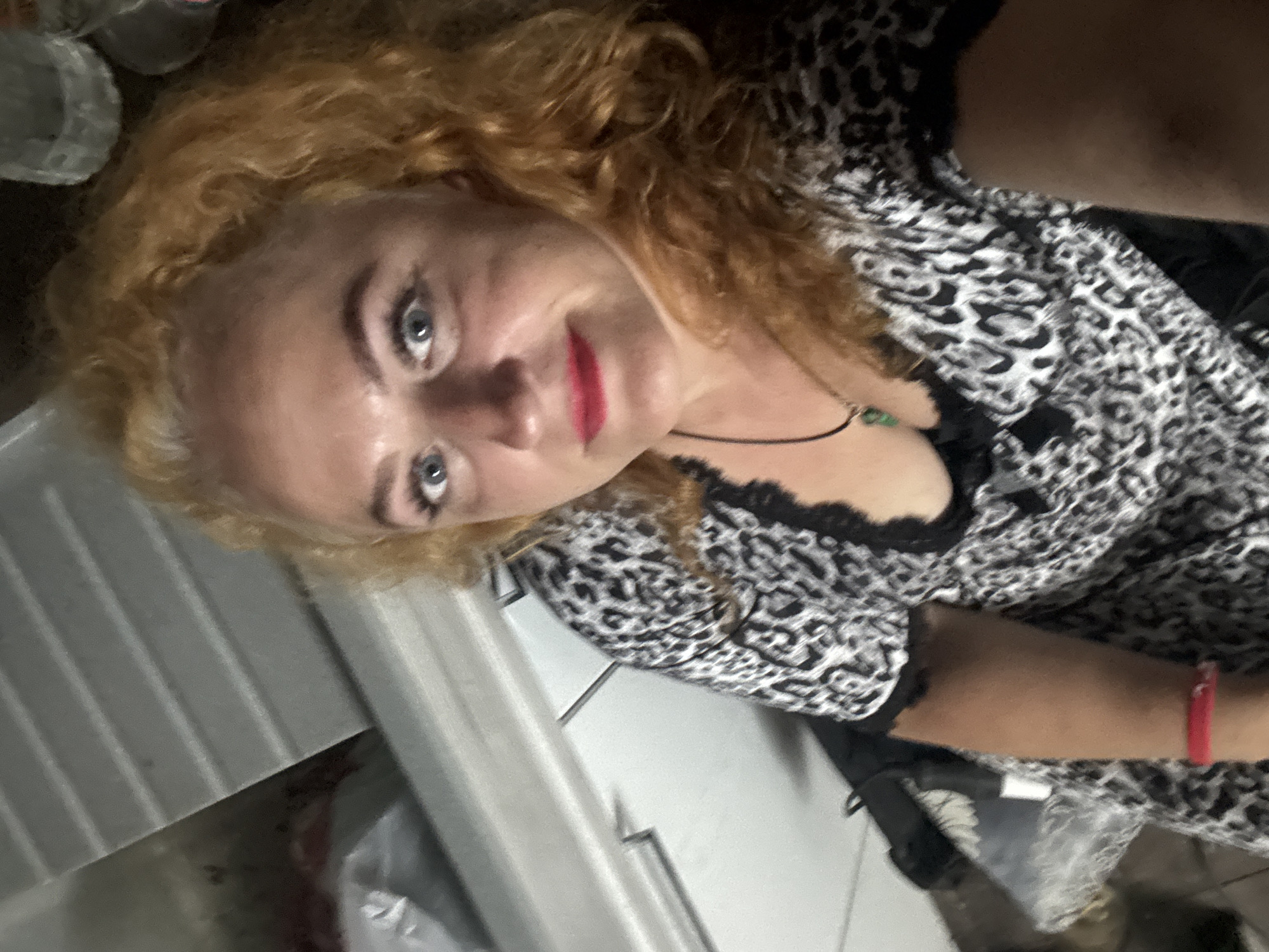 A woman with red curly hair and blue eyes is shown in a selfie. She is wearing a black and white leopard print dress with black lace trim. She has bright red lipstick and a necklace with a green pendant. Her right arm is visible, with a red bracelet on her wrist. In the background, there is a white countertop and what appears to be a metal surface with horizontal grooves. The location is not specified in the image or accompanying information.