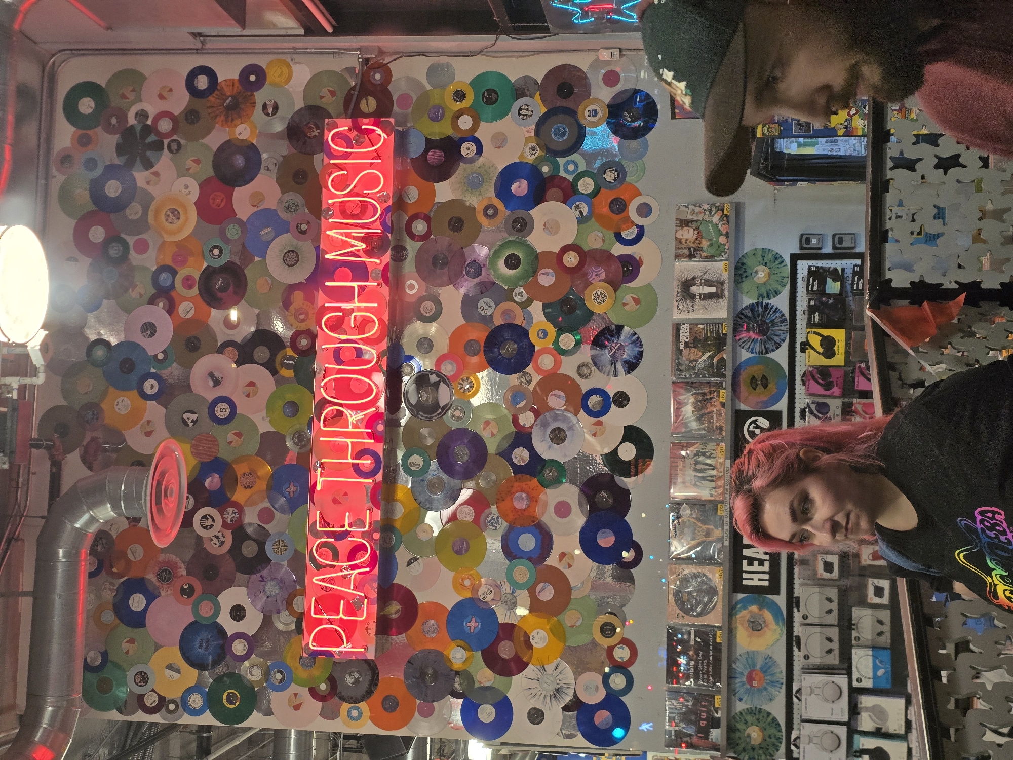 The image shows a store interior with a wall decorated with numerous vinyl records of various colors and sizes. A neon sign above the records reads "PEACE THROUGH MUSIC" in red letters. In the foreground, a woman with pink hair wearing a black t-shirt stands on the left, and the side profile of a man wearing a green baseball cap and a red shirt is visible on the right. Behind them, shelves display records and packaged items. The overall impression is of a music-themed retail environment.