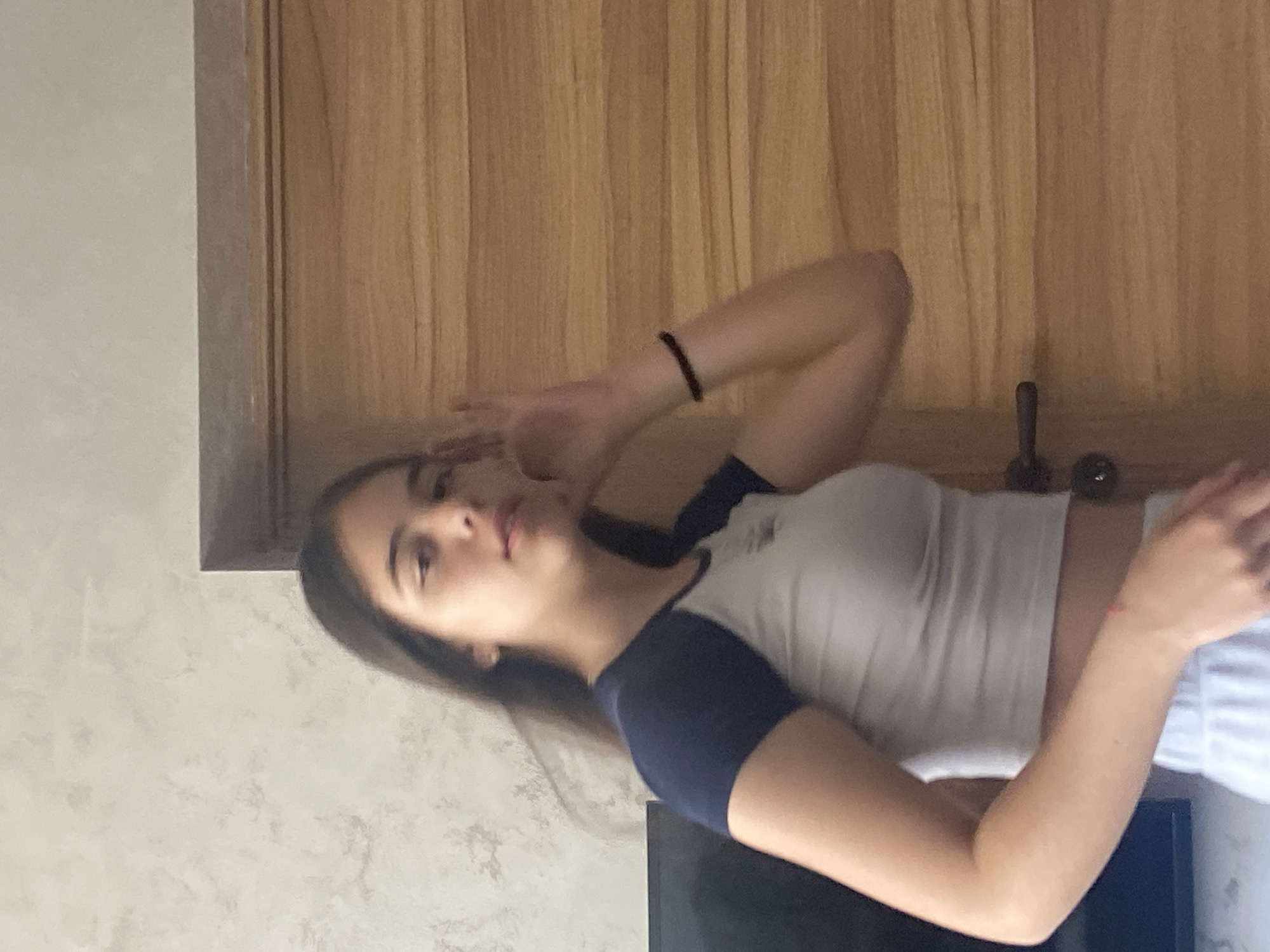 A woman is indoors, posing. She has dark hair and fair skin, and wears a short-sleeved shirt with dark blue sleeves and a white body, along with light-colored bottoms. Her left hand is raised near her forehead. She is positioned in front of a wall painted a light color, with wooden panels and a doorframe visible behind her. A black object, possibly a television screen, is situated near her. The location is unknown. There is no visible indication this image was taken in Dragovishtitsa, Bulgaria.