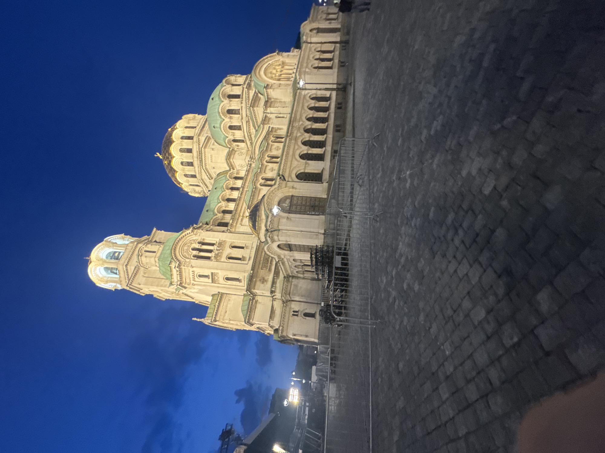 The image captures the Alexander Nevsky Cathedral in Sofia, Bulgaria, at dusk or dawn. The prominent Orthodox cathedral, constructed from light-colored stone, is illuminated, highlighting its ornate architecture, including multiple green-oxidized copper domes and a central golden dome topped with a cross. A tall tower-like structure with a golden cap is visible on the left side of the cathedral. The foreground consists of dark grey cobblestone paving. Several temporary metal crowd control barriers are positioned horizontally across the mid-ground. To the left, a scaffolding structure with stage equipment and lighting fixtures is present, along with an illuminated streetlight. A single, indistinct individual is partially visible walking on the far right side of the frame, near the cathedral. The sky above is a deep blue with darker cloud formations.