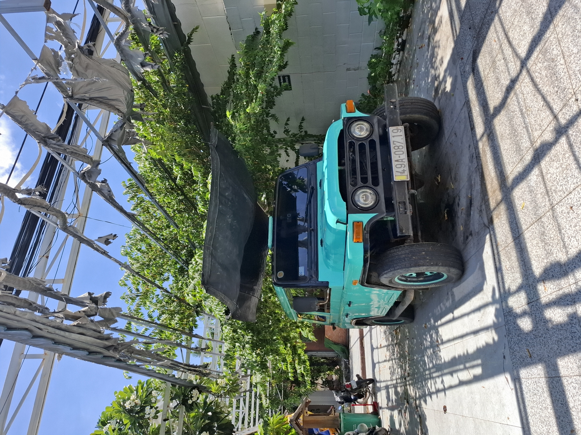 In Phu Thuy Ward, Vietnam, a teal-colored off-road vehicle, possibly a Land Rover Defender, is parked outdoors. The vehicle has a black fabric covering draped over its roof. It is positioned on a paved surface that reflects the dappled sunlight filtering through an overhead structure. This structure consists of a metal frame with torn, grey fabric or sheeting hanging from it, creating a weathered, partially sheltered area.
Behind the vehicle, dense green foliage from trees and bushes grows against a white brick wall. To the left of the vehicle, a motorcycle is parked near a small, ornate shrine-like structure. The ground is a tiled pathway with shadows cast by the overhead structure and foliage, indicating a sunny day. The overall scene suggests an outdoor space that may be a private courtyard or a shaded parking area in a tropical or subtropical climate.