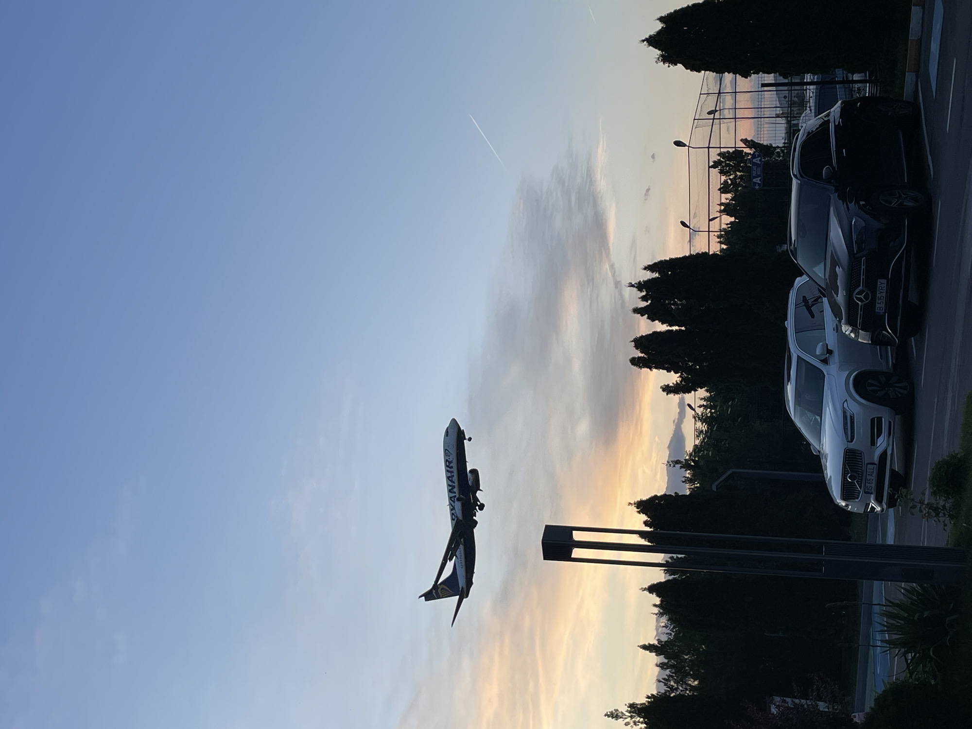 The image shows a Ryanair Boeing 737 aircraft approaching for landing at Henri Coandă International Airport in Otopeni, Romania. The aircraft is seen against a sky with some clouds and a faint contrail. The plane is near the airport perimeter, as indicated by the nearby street lights and trees. Several parked cars are visible on the right, including a black Mercedes-Benz and a white Volvo. A fence and a building can be seen in the background.