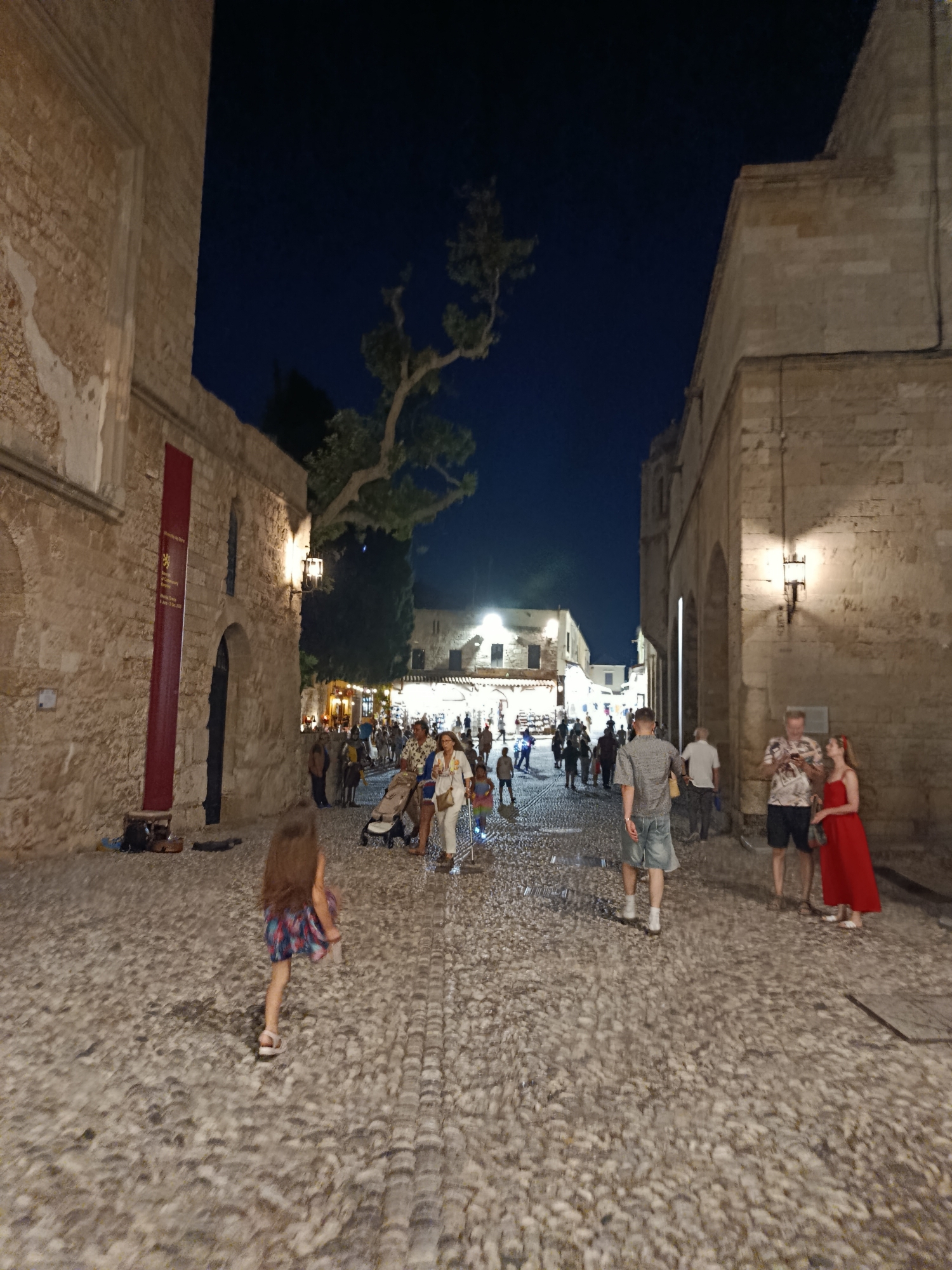 The image depicts a nighttime scene in a cobblestone street, likely in the Municipal Unit of Rhodes, Greece, given the architectural style. Several people are walking along the street, which is lined with stone buildings. Some individuals are standing and talking, while others appear to be browsing shops. A young girl with long brown hair is walking away from the camera towards the right side of the frame. She is wearing a colorful dress and sandals. In the foreground, a man in a plaid shirt and shorts is walking away from the camera. Further down the street, a couple is standing together, the woman in a red dress and the man in a patterned shirt and dark shorts. A baby stroller is also visible. The sky is dark, indicating it is nighttime. The street is illuminated by overhead lights and lamps attached to the buildings, casting a warm glow on the scene. There is a large red banner hanging on the left-hand building with some text on it, though it is not clearly legible. The overall atmosphere appears to be one of a lively evening in a historic European town.