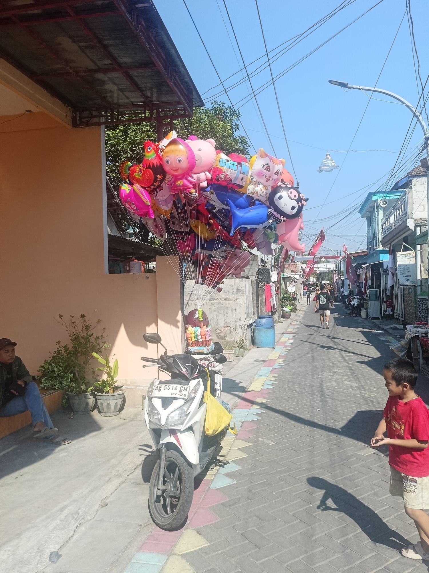 In Gedangan, Indonesia, on a sunny day, a street vendor is positioned next to a parked white motorcycle, displaying a large cluster of colorful cartoon-themed balloons. The balloons feature characters like a rooster, a girl with a pink hat, a bear, a cat, and Hello Kitty. A person wearing a dark jacket and jeans sits on a concrete ledge to the left, near potted plants. To the right of the motorcycle, a young boy in a red t-shirt and khaki shorts stands on a paved walkway, casting a shadow. The street is lined with buildings, some with blue accents, and decorated with red flags. Utility lines crisscross overhead.