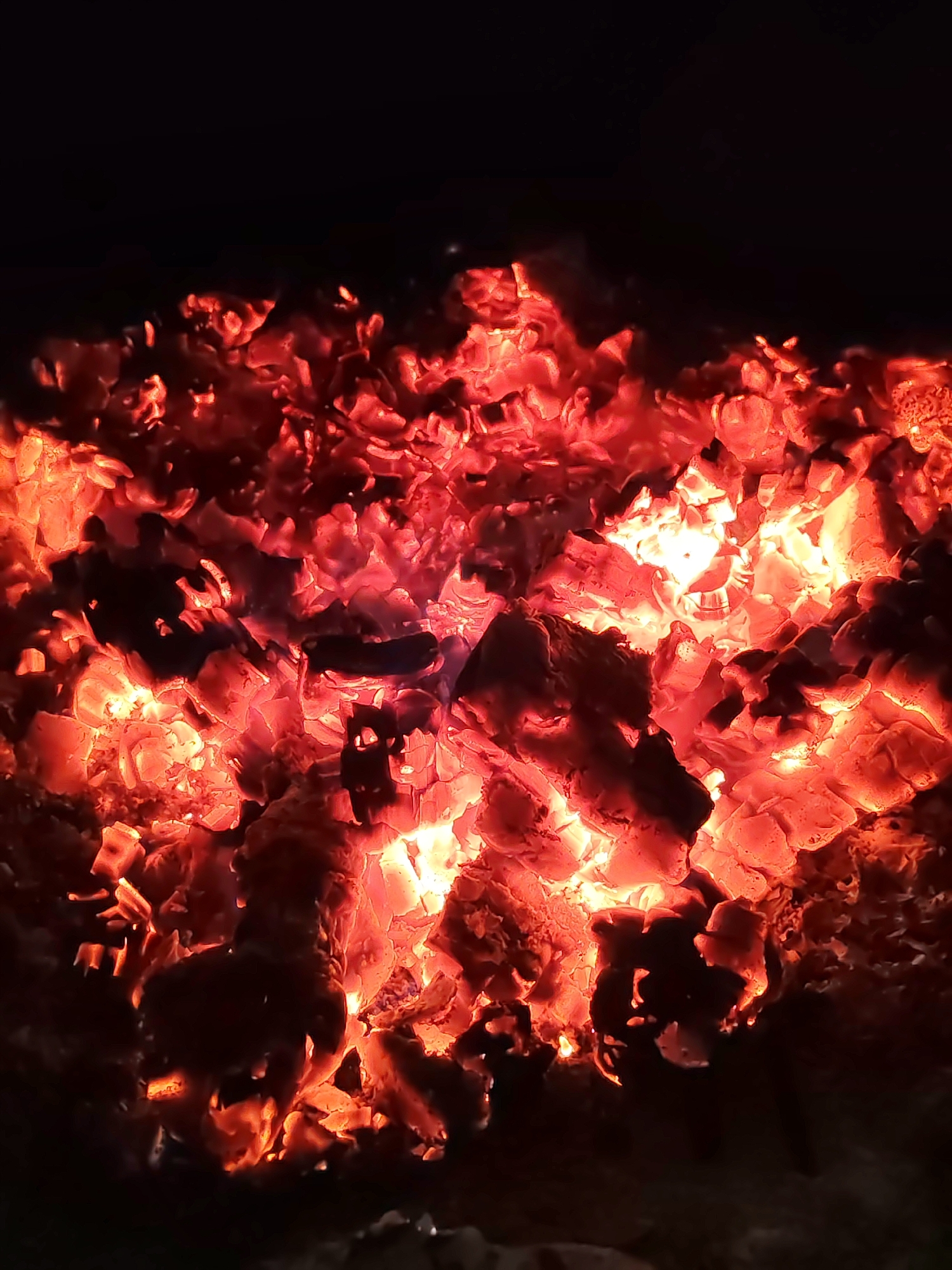 The image shows a close-up view of glowing embers in the dark, possibly in Unknown. The embers are a mix of bright red and orange, filling most of the frame and fading into the darkness above. They appear to be densely packed, creating a fiery texture.