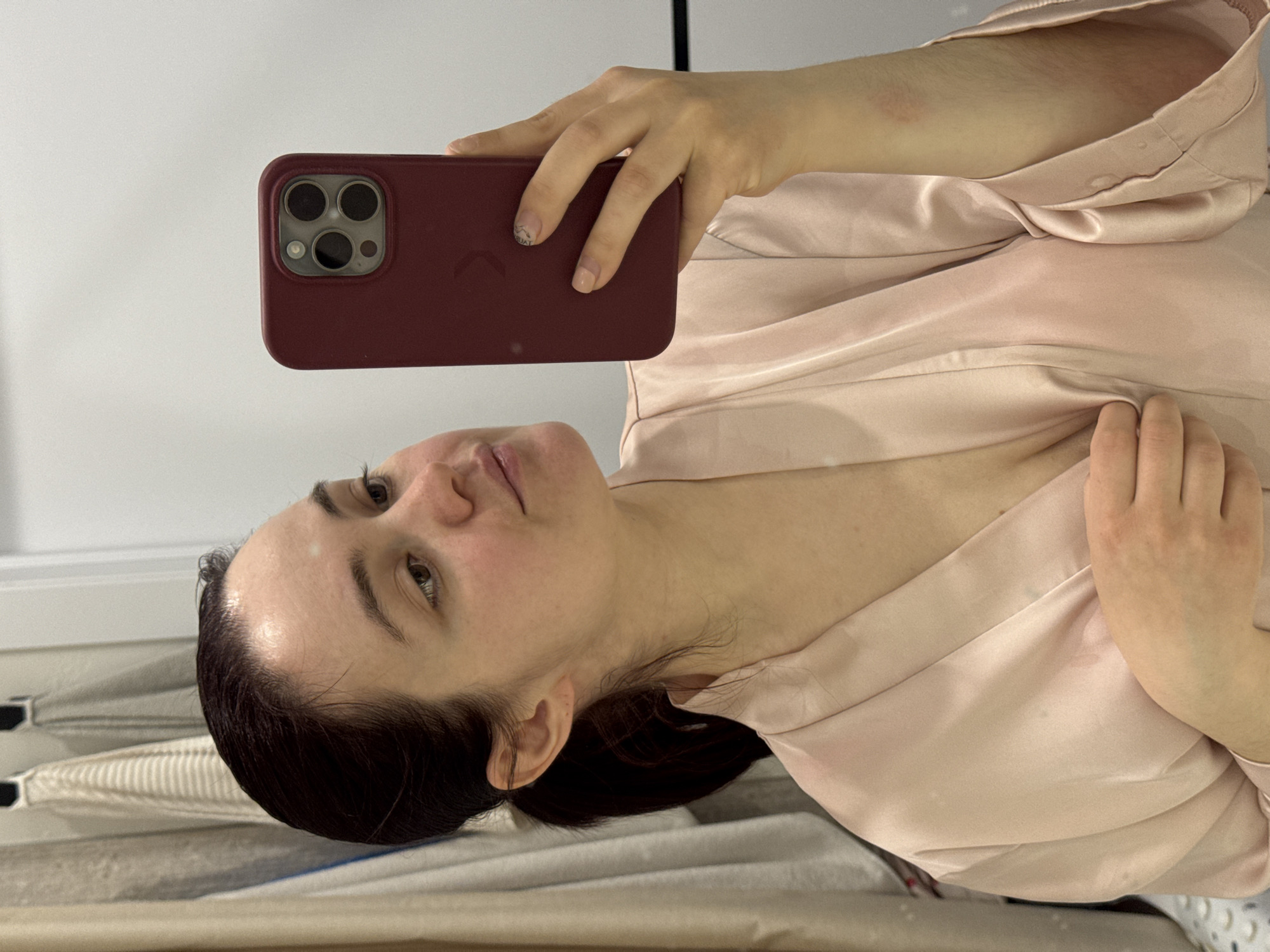 A woman is captured in a mirror selfie, positioned horizontally in the frame. She holds a maroon-colored smartphone with her left hand, its triple-lens camera array clearly visible on the back, suggesting an iPhone model. The woman's head is turned slightly, with her gaze directed towards the camera/reflection. Her expression is neutral, hinting at relaxation or quiet contemplation, with a subtle rosy blush on her cheeks.
She is wearing a light pink or peach-colored satin robe, which appears draped and folded around her torso and arms. Her dark hair is pulled back from her face, revealing her forehead and ear, with some fine baby hairs at the hairline. Her nails are short and natural-looking, with a faint, light-colored design visible on the ring finger of her left hand. A minor reddish mark or irritation is visible on her right upper arm, near the shoulder.
The background primarily consists of light-colored surfaces, including what appears to be a white wall or mirror frame. To the left of her head, there are hints of striped fabric, possibly sheets or another draped item, suggesting an indoor, likely domestic setting such as a bedroom or bathroom. No discernible text is visible anywhere in the image.