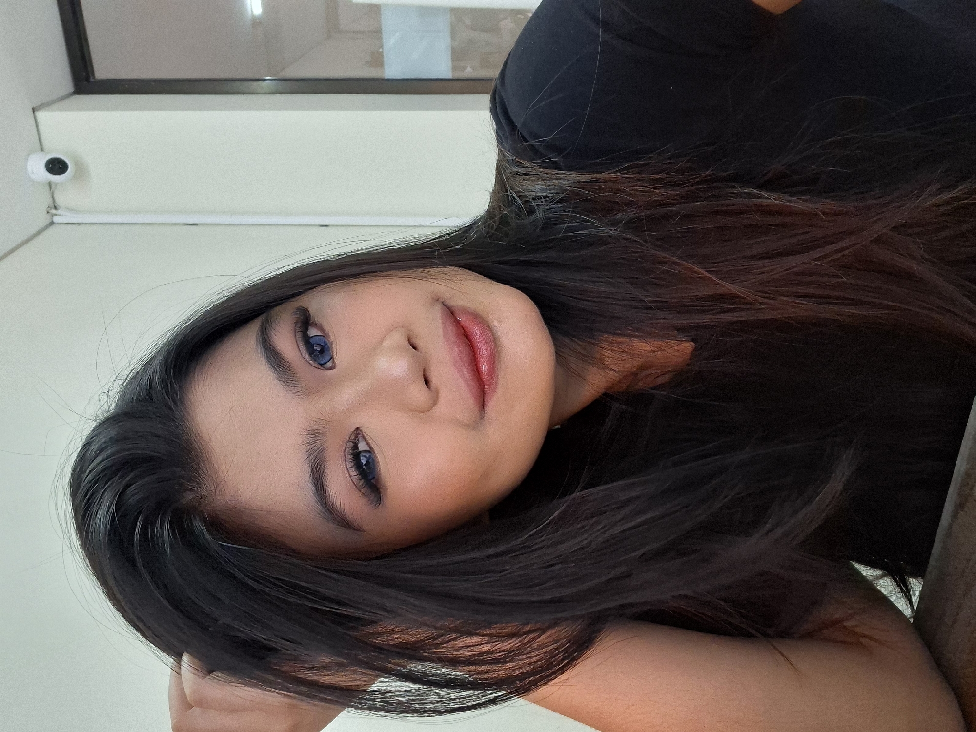 A young woman with dark hair and blue contact lenses is posing for a selfie in what appears to be an indoor setting. She is wearing a black t-shirt and has her left hand in her hair. The background consists of a light-colored wall with a small white security camera mounted near the ceiling in the upper right corner, and a window or glass partition to the right. The scene is likely an office or commercial space in Kuta, Indonesia.