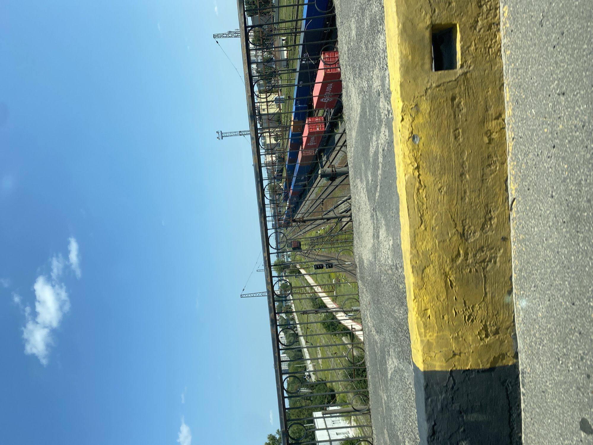 The image captures a view from a bridge overlooking a railway yard. In the foreground, a concrete barrier is visible, painted with a yellow and black hazard stripe. Beyond the barrier, a metal railing with circular openings separates the viewpoint from the railway tracks. Several freight trains are parked on the tracks, some carrying red shipping containers labeled "BOLUDA" and blue containers. The railway yard is surrounded by greenery, and in the distance, buildings and infrastructure typical of a city can be seen, including power lines and tall metal towers. The sky above is clear and blue with scattered white clouds. The scene suggests an urban or industrial setting, consistent with the city of Brest, Belarus, where railway infrastructure is a significant feature.