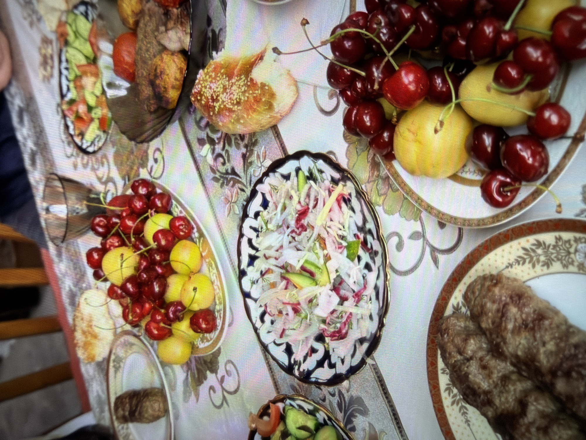 An overhead shot shows a table laden with food, suggesting a meal is in progress. On the left, two grilled meat skewers rest on a decorative plate. To their right, a white oval plate holds a salad composed of thinly sliced vegetables, possibly cabbage and radishes. Further right, a dark bowl contains grilled meat patties alongside red tomatoes. In the foreground, a large plate overflows with dark red cherries and several yellow fruits, possibly plums or apricots. Another plate, further back on the left, displays a small portion of grilled meat. A patterned tablecloth with floral motifs covers the table. The background is blurred but shows wooden chairs and what appears to be a glass. The setting is likely a home or restaurant in Fergana City, Uzbekistan, given the prevalence of such dishes and serving ware in the region.