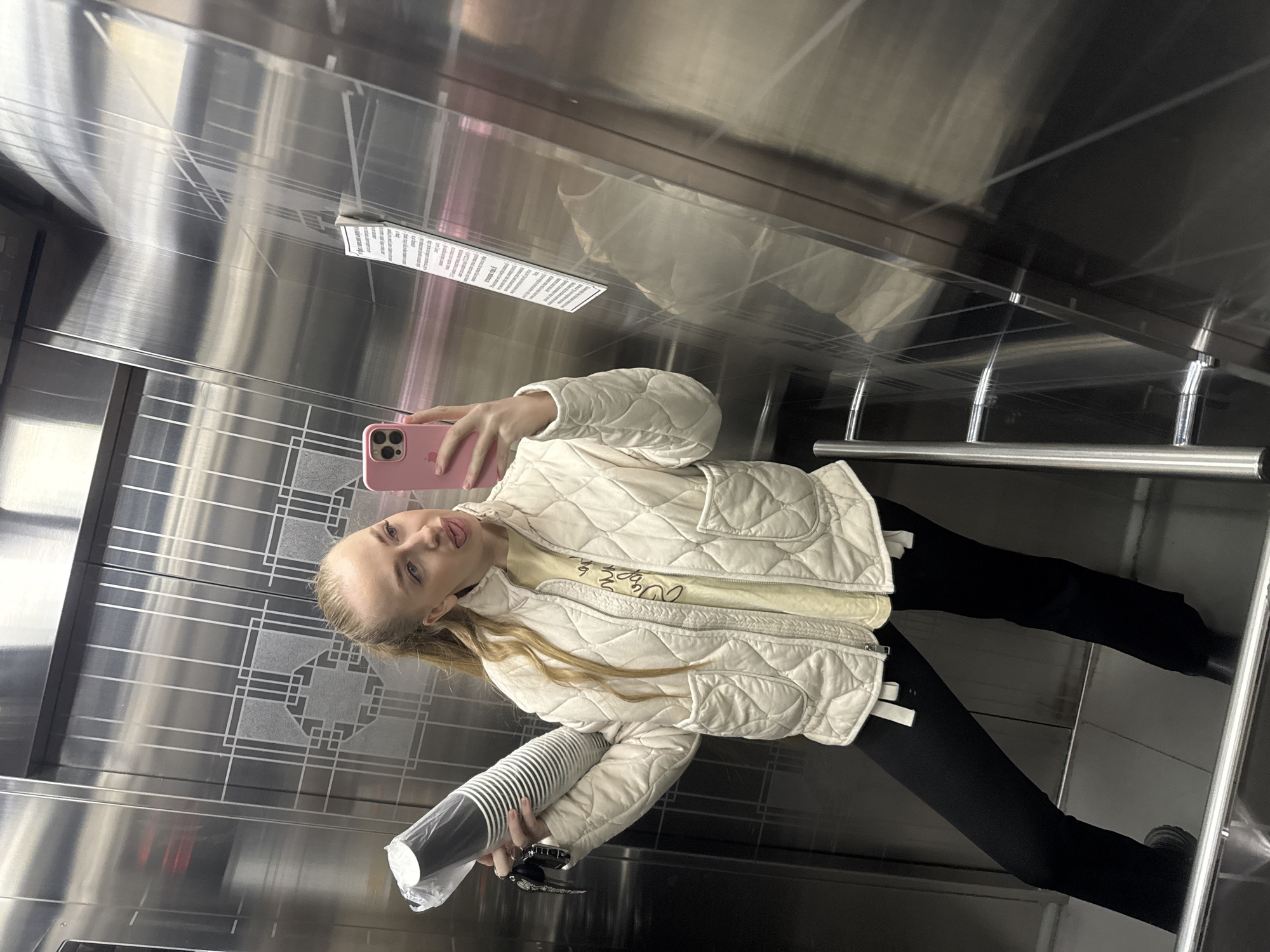 A young woman is taking a selfie in what appears to be an elevator. She is holding a stack of disposable cups in one hand and a pink smartphone in the other. She is wearing a white quilted jacket, a light yellow top, and black leggings. The elevator has brushed metal walls with a geometric pattern. The scene is enclosed, with metallic bars visible on one side. There is a sign or label affixed to the wall above her. The location of Petropavlovsk-Kamchatsky, Russia is not verifiable from the image.