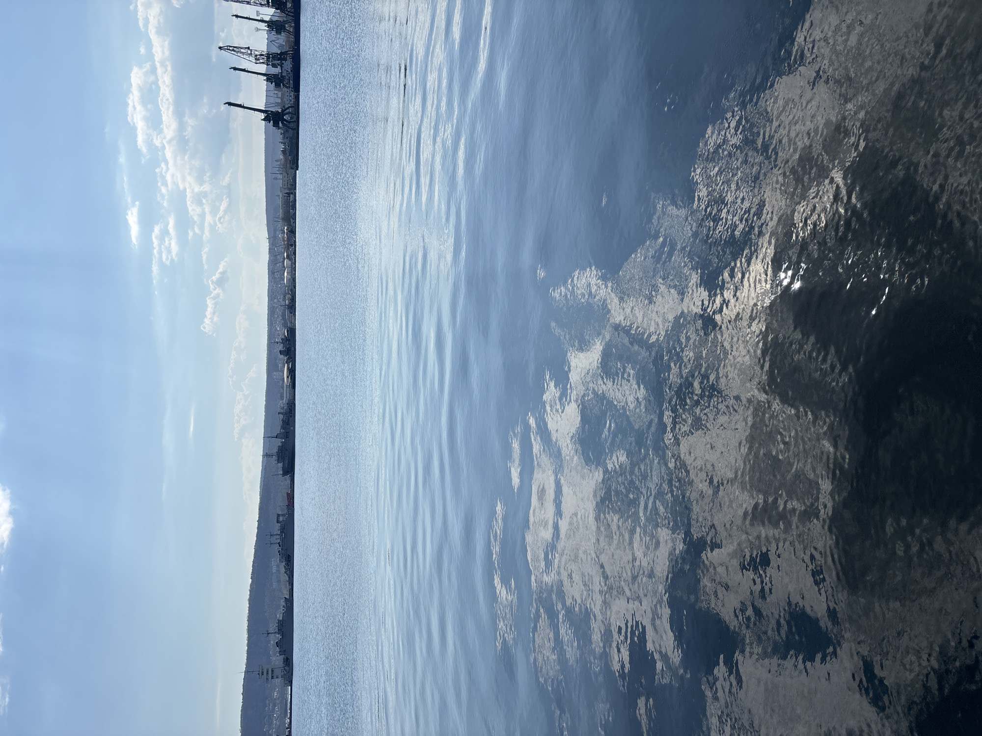 The image captures a view of the sea in Varna, Bulgaria, with the city's port infrastructure visible along the coastline. Cranes and buildings are silhouetted against a sky with scattered clouds. The sea occupies the majority of the frame, with variations in color and texture indicating differing depths and light reflections. The angle of the photo appears to be taken from a tilted point of view or a boat.