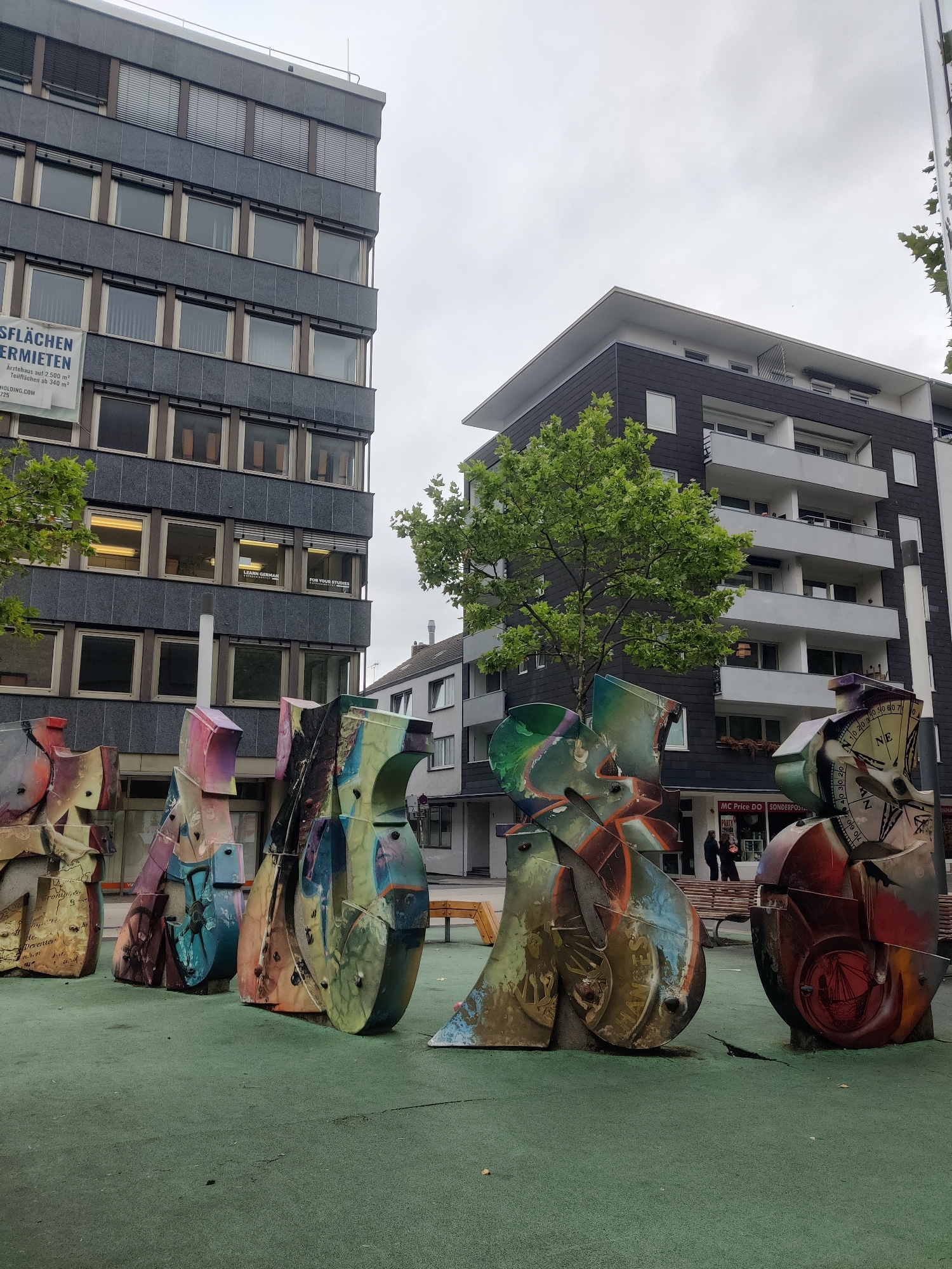 The image shows a row of colorful, abstract sculptures in an urban setting. The sculptures are made of metal and appear to be abstract representations of figures or objects, with vibrant paint and varied textures. They are arranged on a green, textured surface, possibly a park or a public square.
In the background, two buildings are visible. The building on the left is a multi-story structure with a dark, grey facade and numerous windows. A banner with German text, "FLÄCHEN ZU VERMIETEN" (Spaces for Rent), is attached to its side. The building on the right is a modern apartment building with a dark facade, white balconies, and large windows. Trees with lush green foliage are present, partially obscuring the buildings.
In the middle ground, a few people can be seen walking near a shop entrance. A wooden bench is also visible. The sky is overcast, suggesting a cloudy day.
This scene is located in Dortmund, Germany. Specifically, this public art installation is situated in front of the city library and the former Commerzbank building at Reinoldikirchplatz, which is in the city center of Dortmund.