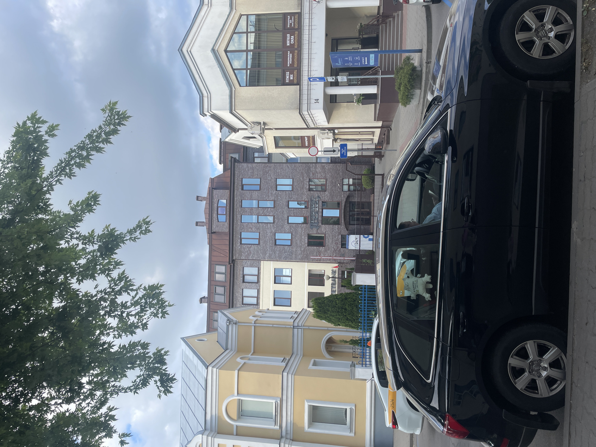 This detailed image depicts an urban street scene in Brest, Belarus, specifically around Sovetskaya Street (ул. Советская), with building numbers 12 and 14 clearly identifiable. The setting is under a cloudy sky, with dense green tree foliage partially obscuring the upper portions of the buildings.
From left to right, the buildings include:
On the far left, a two-story building with a light yellow facade, white decorative trim, arched windows and doorways, and a light gray pitched roof. A blue metal fence with white posts runs along its front.
Adjacent to it, a multi-story building with a lighter cream-colored facade, brown-framed windows, and a partially visible "KEYBANK" sign. A small red and white flag is displayed near its entrance.
Behind these, a prominent six-story building constructed of dark reddish-brown brick features numerous rectangular windows with dark frames and a dark brown sloped roof. A black metal gate provides access to a property in front of this building.
On the right, a two-story cream-colored building, marked with the number "14" on its facade, presents a large, reflective central window on its upper floor. This window displays white text, including "FITNESS MIX," "ФУНКЦИОНАЛЬНЫЕ ТРЕНИРОВКИ" (Functional Training), "МЯГКАЯ СИЛА" (Soft Power), and "ТАНЦЕВАЛЬНЫЕ ЗАЛЫ" (Dance Halls), along with the repeating name "ПАЛАУМ" (Palaum). The building has a symmetrical entrance with steps, two columns, and planters. A white sign with "РЕФОРМЕР" (Reformer) is affixed to its side.
The foreground is dominated by a paved street and sidewalk. A dark-colored (likely black) Audi Q5 SUV is parked curbside, facing right, with its driver visible inside, looking down at an object (possibly a phone or book). A white, cartoon-like plush toy hangs in the SUV's rear passenger window. Behind the Audi, a white sedan, possibly a taxi (indicated by a yellow stripe on its roof and the number "2524" on its side), is also parked.
Street furniture includes a blue parking sign with a white 'P' and an arrow, explicitly indicating "ул. Советская, 12" (Sovetskaya Street, 12) and a date "10.05.20." Other signs visible are a circular red traffic sign (likely a "no entry" or "no stopping" sign) and a blue rectangular sign with a white downward-pointing arrow. The overall scene conveys a typical urban environment with commercial and residential structures, parked vehicles, and active street signage.