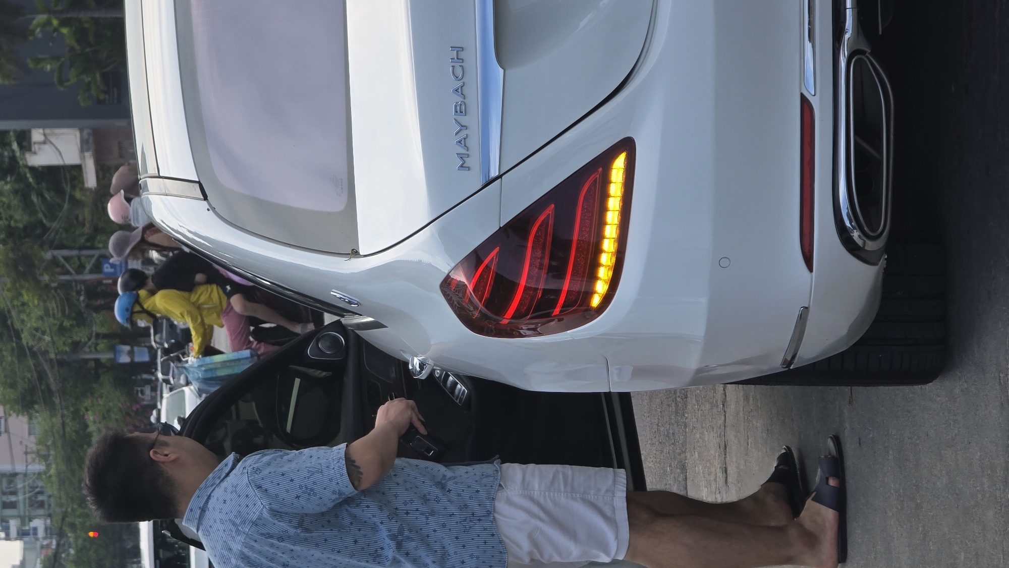 The image displays a sideways view of an urban street scene in what appears to be Nha Trang Ward, Vietnam, during daytime. A white luxury Maybach car is prominently featured, viewed from its rear-right side. The car's distinctive red LED taillights are illuminated, with the right turn signal actively flashing yellow. The "MAYBACH" emblem is clearly visible on the trunk lid, above a chrome accent strip, and a large, chrome-trimmed exhaust outlet is integrated into the right side of the rear bumper.
A man, wearing a light blue patterned short-sleeve shirt, white shorts, and dark sandals, stands by the open rear passenger door of the Maybach. His left arm, adorned with a tattoo, is visible as he leans into the car, suggesting he is either entering or exiting the vehicle.
In the background, several individuals are gathered, some wearing hats and helmets, indicative of a typical street environment in Vietnam. A person in a yellow jacket and blue helmet is visible next to a light blue crate, possibly near motorcycles or scooters. A parking sign with a blue "P" symbol is discernible in the mid-ground. Faintly visible in the far background are trees, buildings, and a red traffic light. The scene is brightly lit, suggesting clear weather conditions.