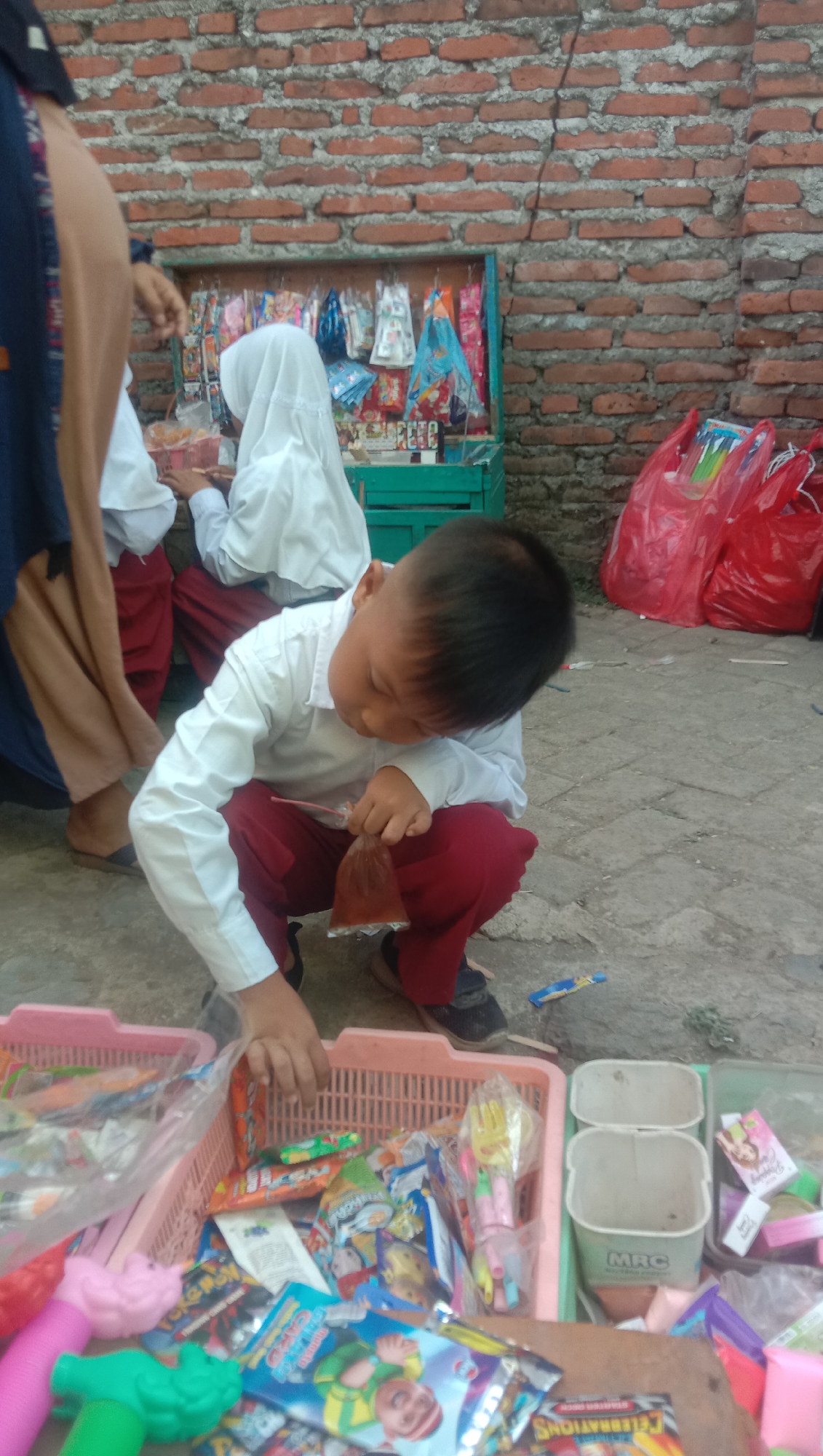 A young boy in a white shirt and red pants is crouching and examining a small plastic bag containing a brown liquid. He is in front of a pink basket filled with various snacks and toys. Behind him, two other children wearing similar school uniforms are partially visible, as well as a stall with assorted small items hanging. The ground appears to be paved, and a brick wall forms the background. The scene suggests an outdoor setting, possibly near a school or a market area in Karang Anyar, Indonesia. Red plastic bags are visible on the right side of the image.