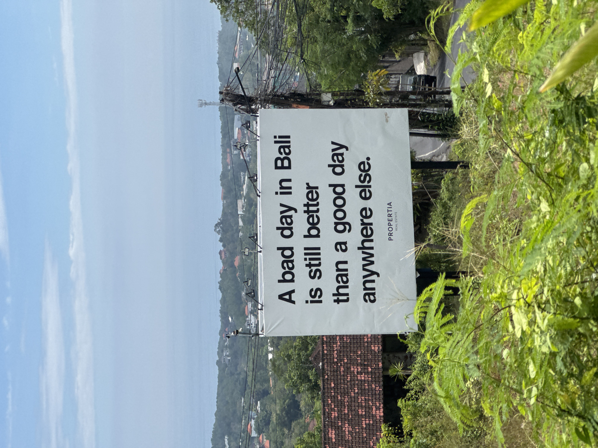 A sign in Kuta Selatan, Indonesia, reads: "A bad day in Bali is still better than a good day anywhere else." The sign, advertising PROPERTIA REAL ESTATE, is mounted on a utility pole amidst lush greenery. A tiled roof is visible below, and in the distance, houses are nestled in the hills near the coast. The sky is pale blue.