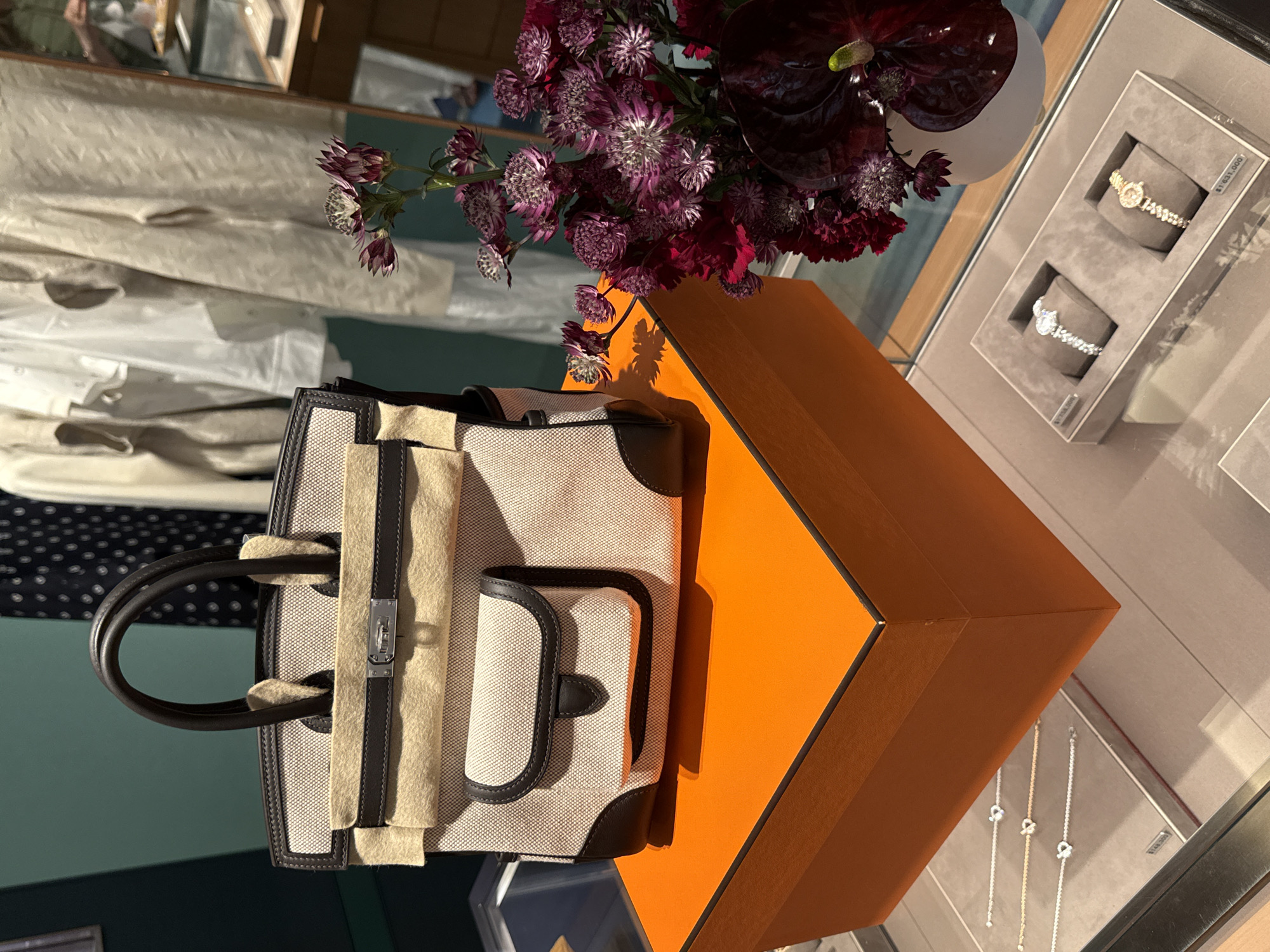 The image shows a still life composition on a glass display surface. A beige tote bag with brown leather trim sits on an orange box. A bouquet of dark purple flowers rests next to the box. A display case with jewelry, including bracelets and watches, is visible in the background. The location is possibly a retail store in Zapopan, Mexico, given the merchandise and the presentation.