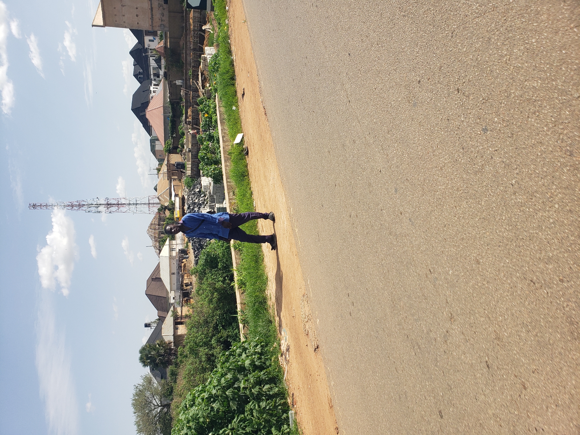 A man in a blue shirt and dark pants walks along a dirt path beside a paved road. In the background, there are several houses with modern roofing, a tall telecommunications tower, and some construction activity. Lush green vegetation lines the dirt path. The sky is clear with scattered white clouds. The scene appears to be in a suburban or developing area.