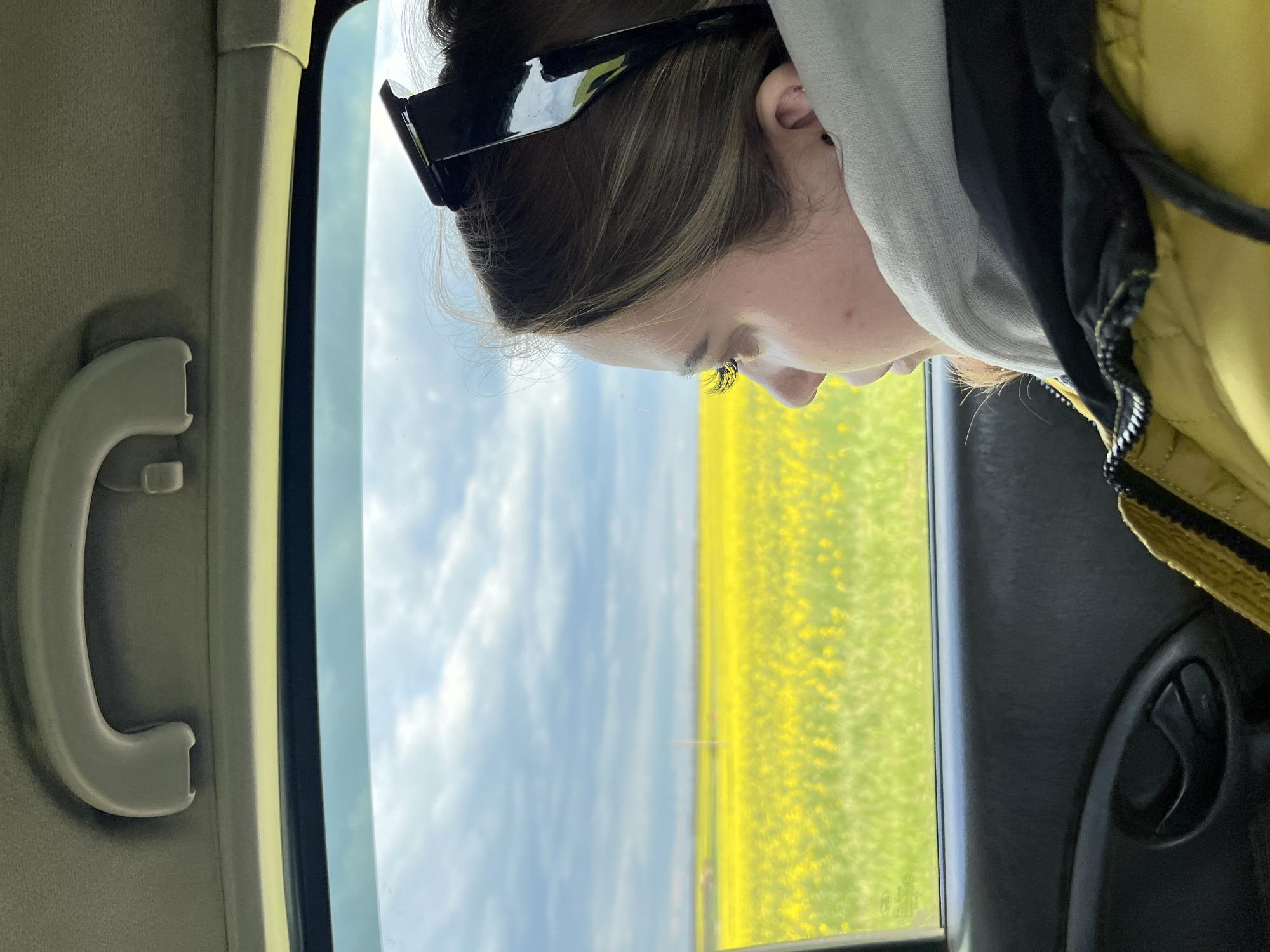 A young woman with sunglasses perched on her head is seen through the open window of a vehicle, likely a car, as it travels through Novomargaritovo, Russia. Outside the window, a field of yellow flowers stretches out under a sky with light clouds. Inside the car, elements of the interior are visible, including the grab handle on the roof near the window and the dark interior of the door panel. The focus is primarily on the woman's profile and the landscape visible through the window.