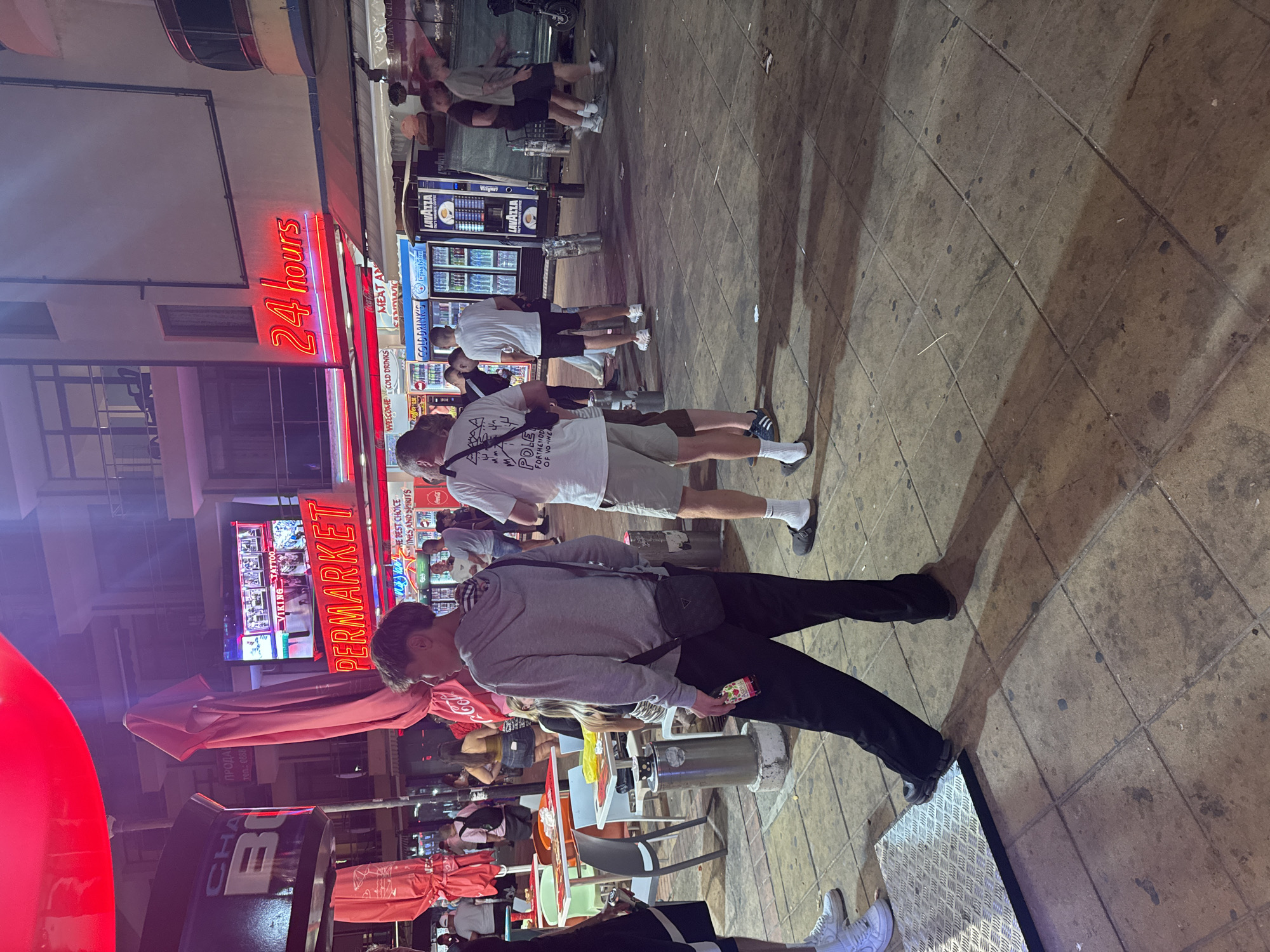 The image depicts a street scene at night in Nesebar, Bulgaria. A brightly lit "PERMARKET" sign is prominently displayed, along with a neon "24 hours" sign. People are standing and walking on a paved area. In the foreground, a young man in a grey hoodie and black pants is holding a small bottle. To his right, a man in a white t-shirt and khaki shorts is also standing. In the background, several other individuals are visible, some near a coffee machine and a row of refrigerators. Umbrellas and tables from an outdoor seating area are also present. The overall atmosphere suggests a bustling commercial area at night.