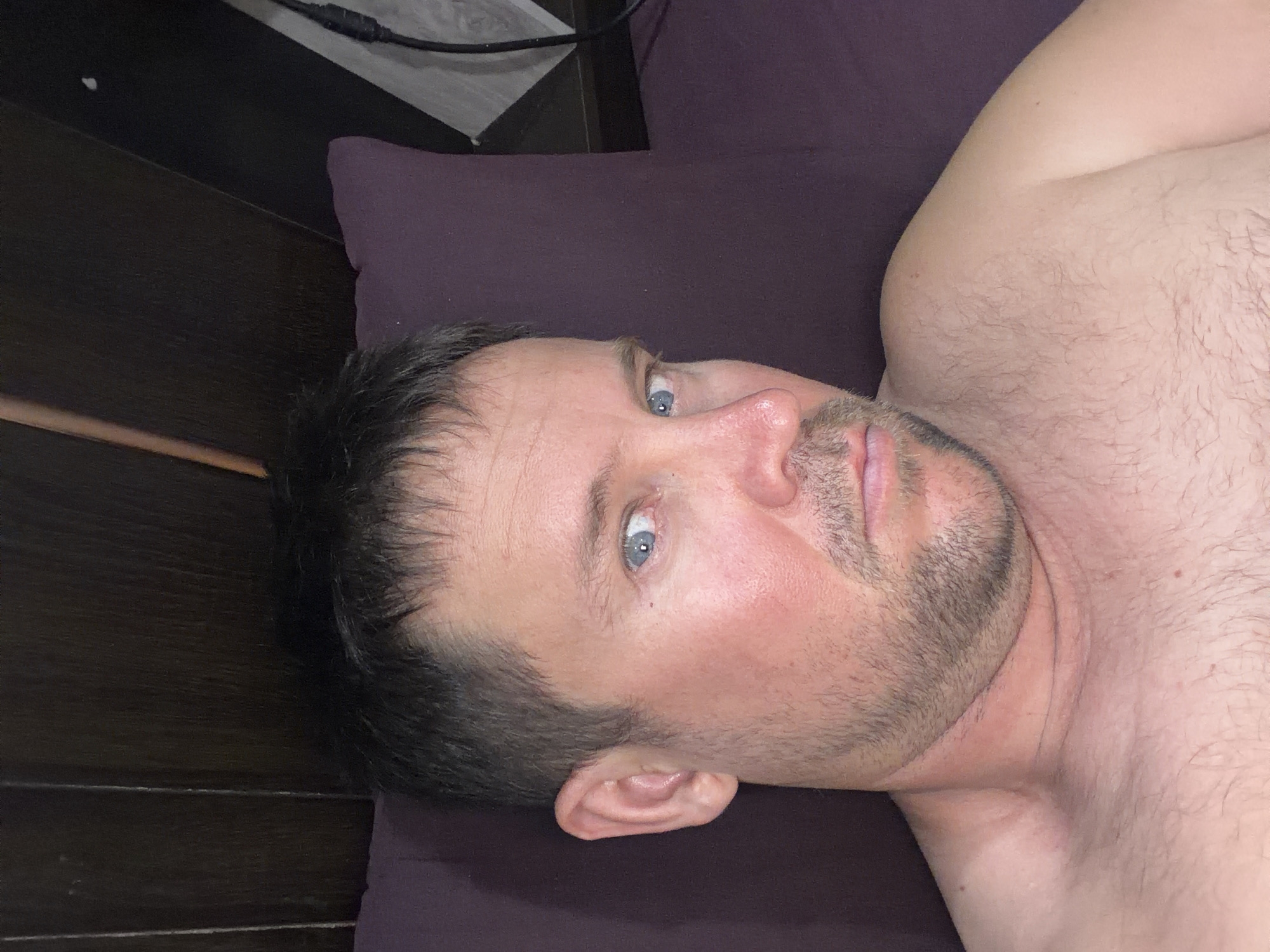 A medium shot, taken from a slightly low angle, captures a man lying on his side in bed. He is shirtless, with dark hair and blue eyes. The man is looking directly at the camera with a neutral expression. He has stubble on his chin and a light beard. The background consists of a dark wooden headboard and a purple pillow. A power cord is visible in the upper right corner of the image. The scene is indoors, likely a bedroom, in Baan Mae Nam, Thailand.