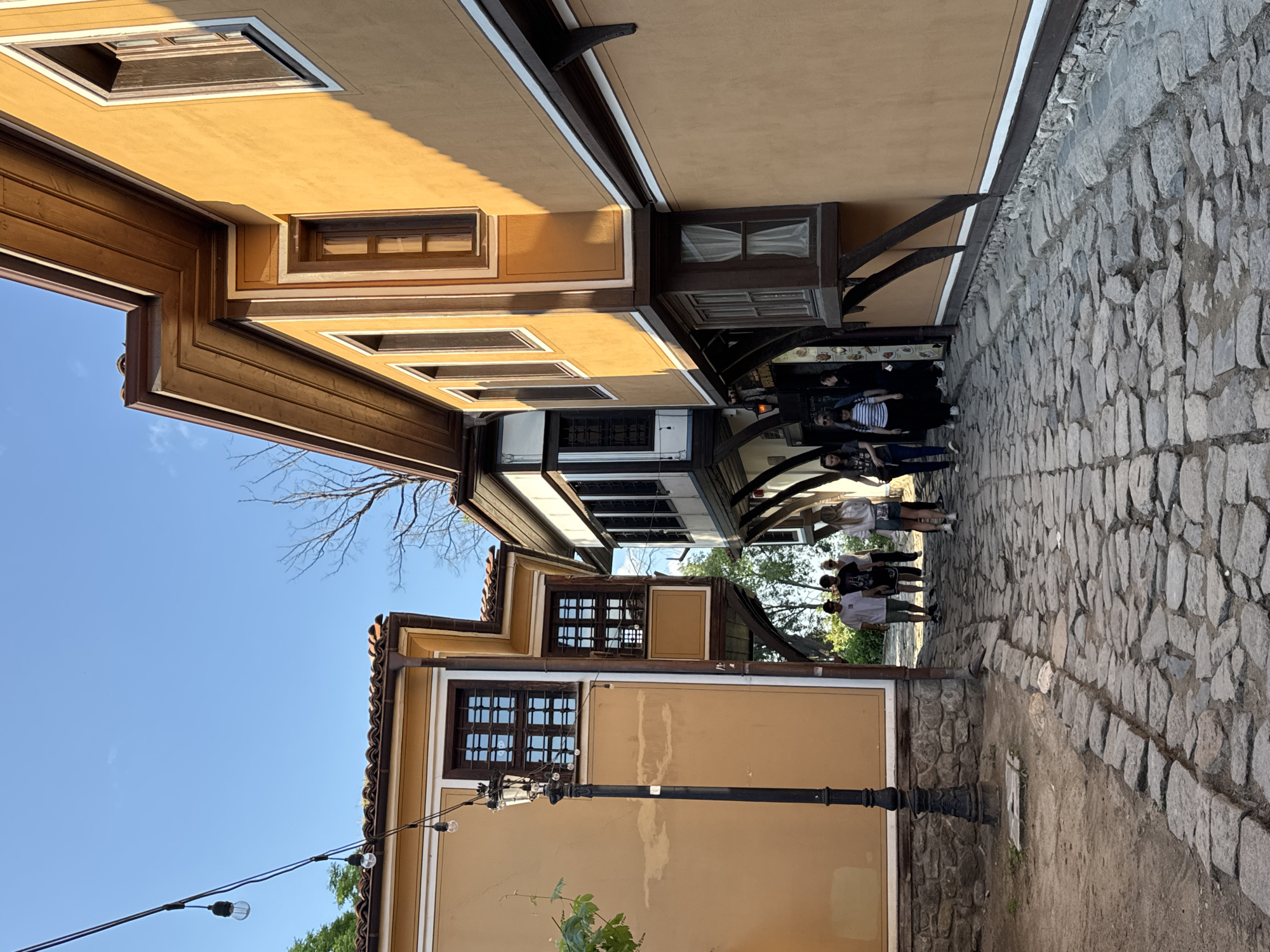 This street scene, likely in the Old Town of Plovdiv, Bulgaria, features buildings with distinctive architectural elements like overhanging upper stories supported by wooden beams, and windows with decorative wooden frames. The buildings are painted in shades of yellow and beige. A cobblestone street runs between the buildings. A small group of people, mostly wearing casual clothing, are standing along the street. A black lamppost stands at the front. A bare tree branch is visible in the upper left of the frame against a blue sky. String lights are also suspended, adding to the ambient lighting of the street.