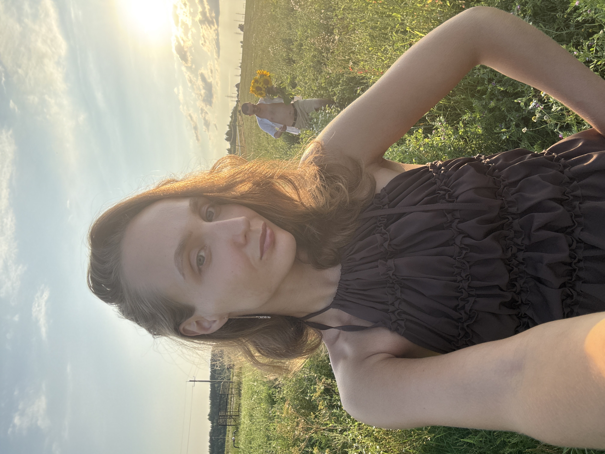 A young woman in a brown ruffled dress is taking a selfie in a field of tall grass and wildflowers, with a man in the background holding a large bouquet of sunflowers. The sun is low in the sky, casting a warm glow over the scene. The woman's hair is light brown and wavy, and she is wearing a long, delicate earring. Her expression is neutral as she looks at the camera. The man is wearing a light blue unbuttoned shirt and tan shorts. He appears to be walking towards the woman. The landscape is rural, with power lines and trees visible in the distance. The location is identified as Aksaitovo, Russia.