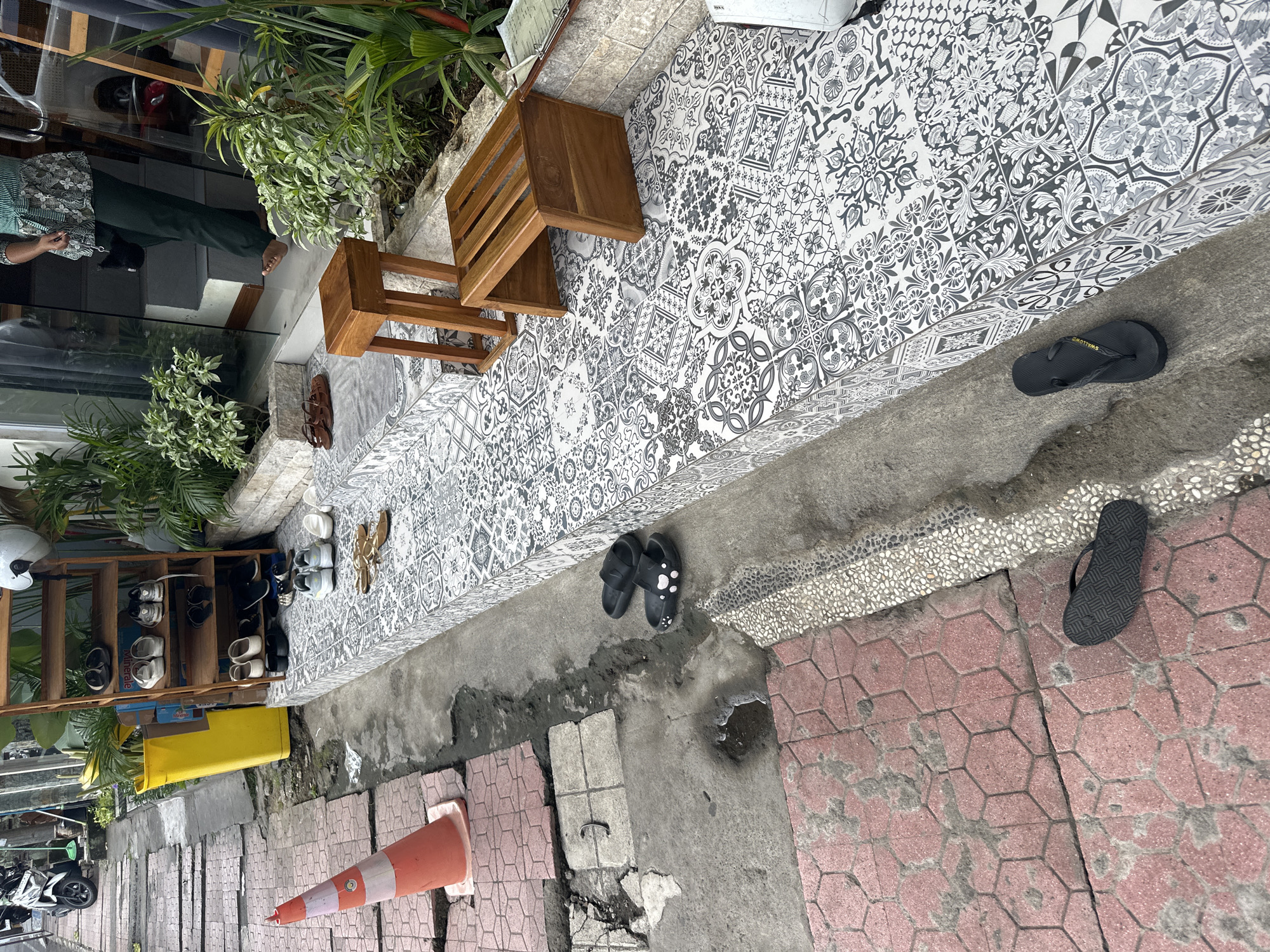 A scene captures the exterior of a building in Ubud District, Indonesia, showcasing a pathway paved with hexagonal red bricks and a tiled step with a black and white mosaic pattern. Several pairs of footwear are visible: black flip-flops with a paw print design rest on the concrete, while a single black flip-flop with a patterned sole lies on the red bricks. A wooden shoe rack stands against the building, holding various pairs of shoes and sandals. A bright yellow plastic bin is positioned next to the shoe rack, and a traffic cone with orange and white stripes is placed on the pathway. A wooden bench-like stool sits on the tiled step. In the background, lush green plants surround the entrance, and a portion of a motorcycle is visible on the left. A person, partially visible in the doorway, is dressed in a green and white patterned top and dark trousers, with one bare foot stepping onto the tiled area.