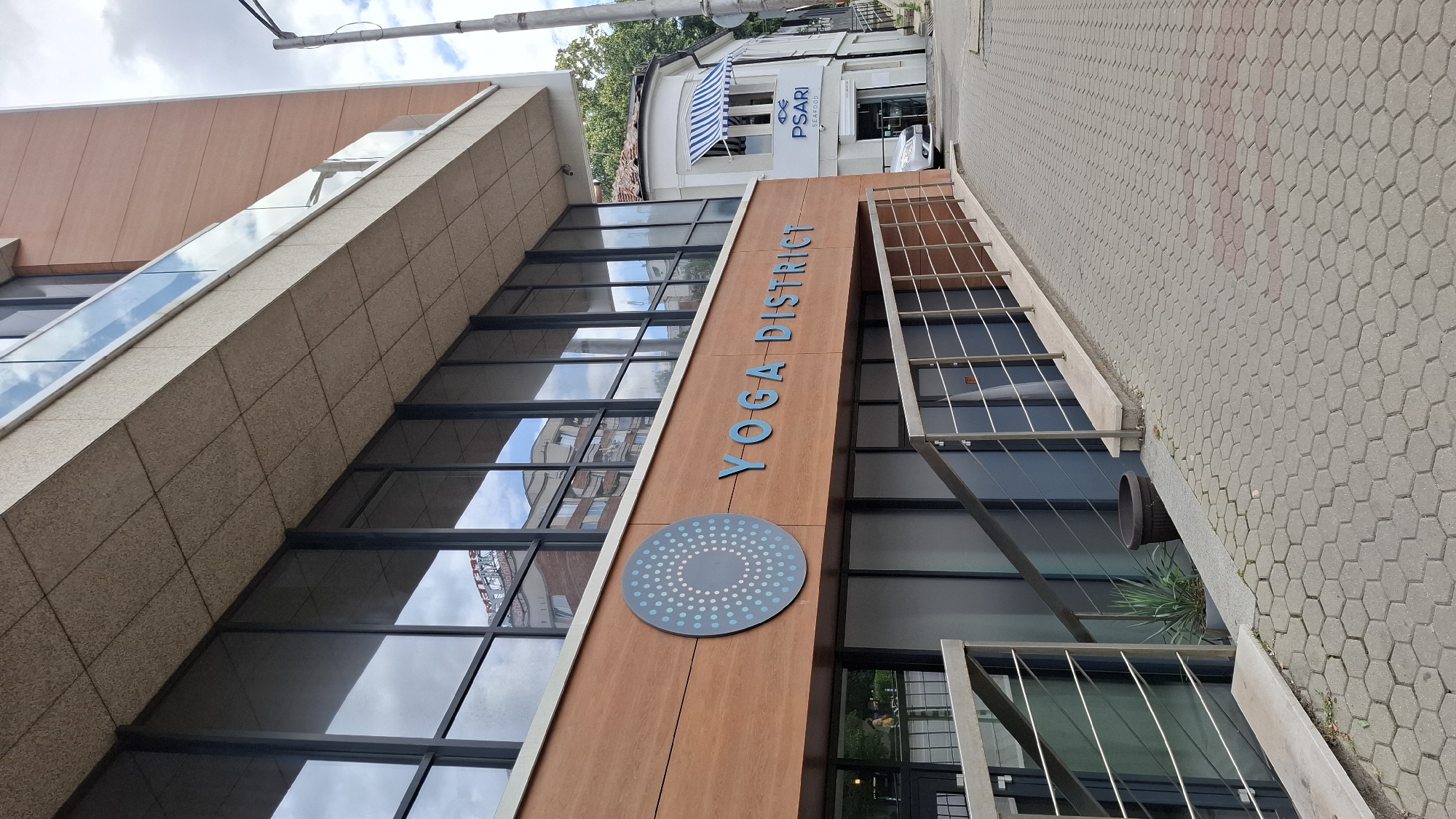 The image shows the exterior of a building in Sofia, Bulgaria, featuring the "YOGA DISTRICT" signage. The building has large windows reflecting the sky and surrounding environment. To the right, a smaller building with blue and white striped awnings displays the sign "PSARI SEAFOOD". The foreground consists of a patterned paved walkway with metal railings. A potted plant is visible near the entrance of the Yoga District. A silver car is parked in front of the seafood restaurant. The sky is partly cloudy.